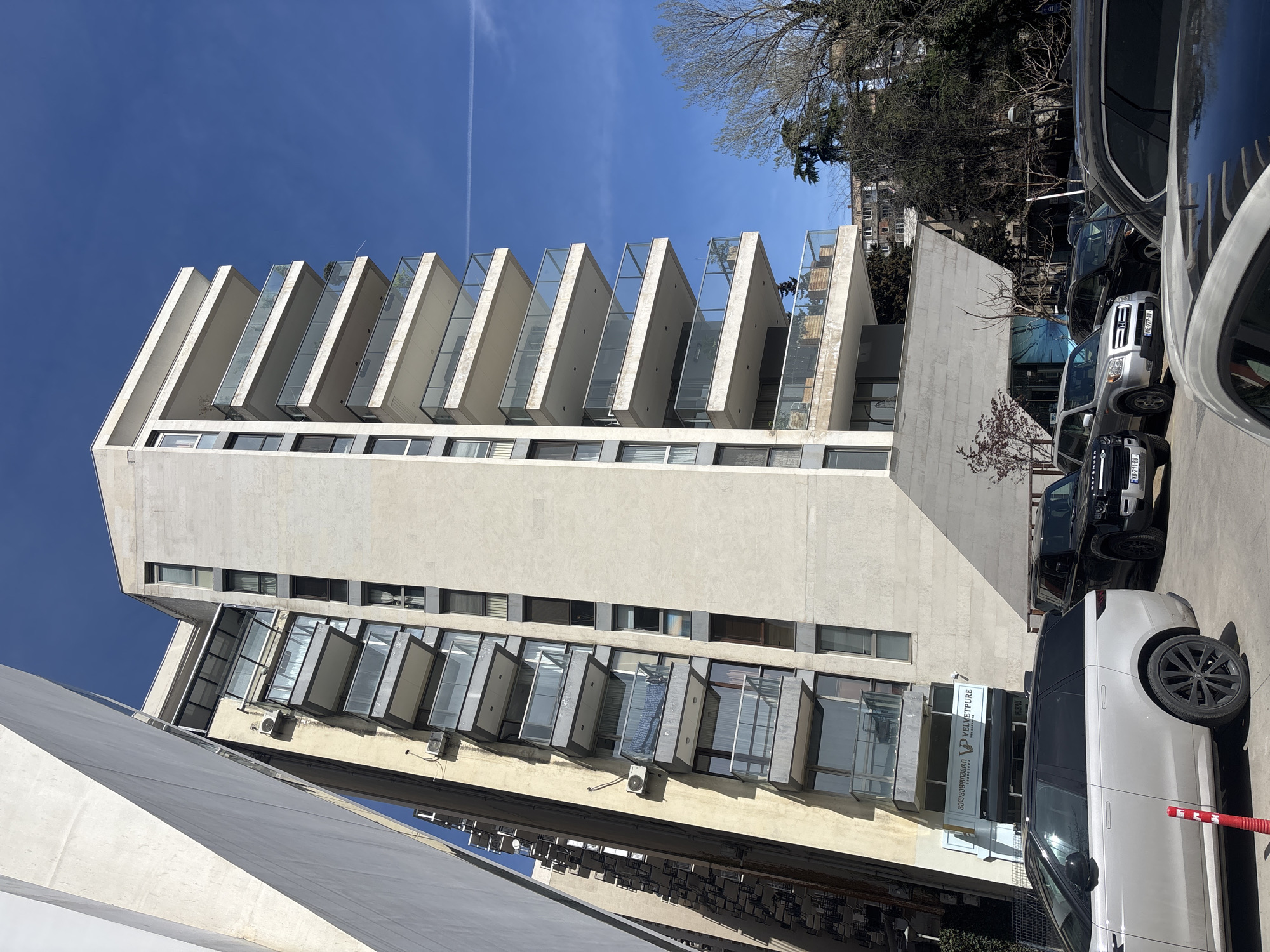 The image captures a tall, modern building with a distinctive architectural design, featuring projecting balconies with glass railings. The building is likely a residential or commercial structure. The sky above is clear and blue, with a faint contrail visible.
Several parked cars are visible in the foreground, indicating an urban environment. A sign "Velvetpure medical clinic" is posted near the building's entrance.
The location is in the city of Tbilisi, Georgia. This is based on Georgian text on the sign, and Georgian license plates.