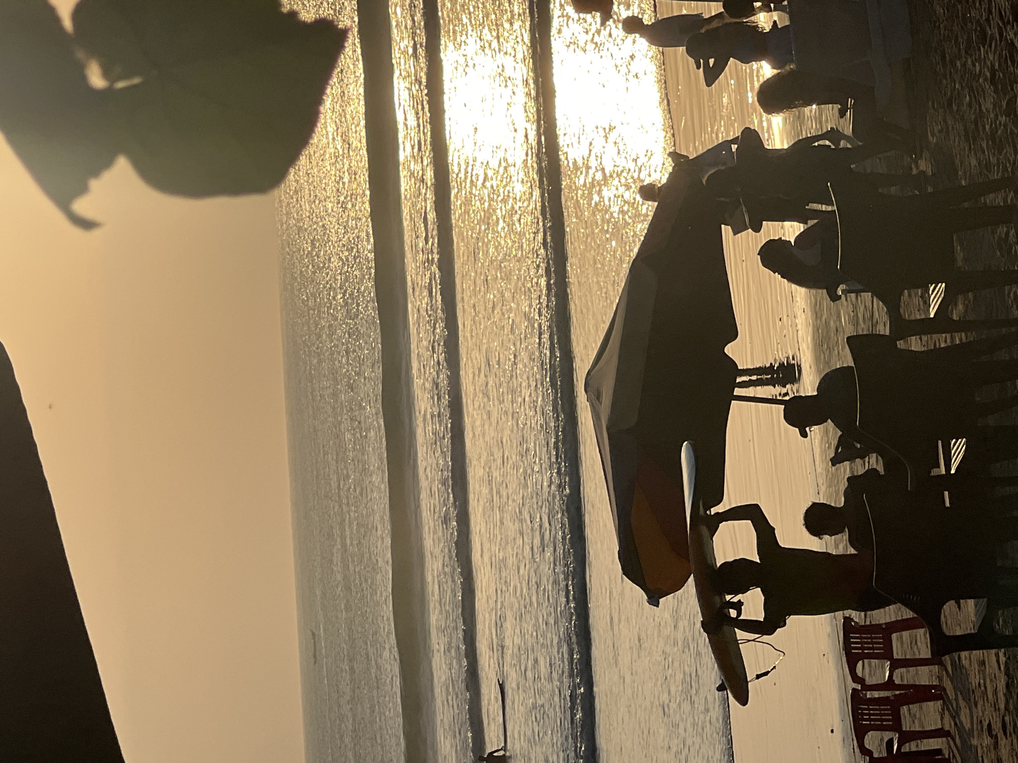 This is a photograph of Kuta Beach in Kuta, Indonesia, at sunset. The sky is a warm orange, and the sun reflects off the ocean, creating a shimmering golden surface. Gentle waves roll onto the sandy shore. In the foreground, several people are silhouetted against the bright water. One person is holding a surfboard above their head. There are beach umbrellas and chairs visible on the sand, suggesting a relaxed atmosphere. Further out in the ocean, a small boat can be seen. The image captures a serene moment on a popular tourist beach.