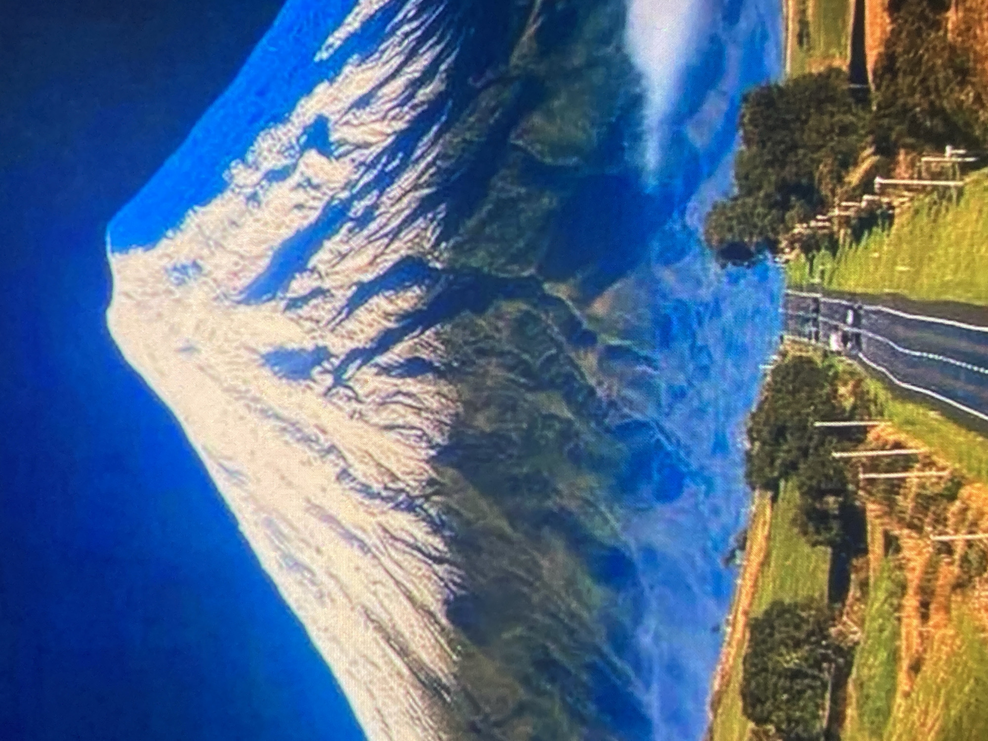 This image depicts a vast, snow-capped mountain dominating the background under a deep blue sky. The mountain's slopes are rugged and appear to be covered in a mix of snow and dark vegetation. A band of blueish mist or low-lying clouds is visible at the base of the mountain. In the foreground, a paved road with white lines curves into the distance, flanked by green fields and scattered trees. There are a few vehicles on the road. The overall scene is scenic and appears to be a rural or natural landscape. The location is identified as Ustyvytsia, Ukraine, suggesting this is a view of a mountain within or near that city.