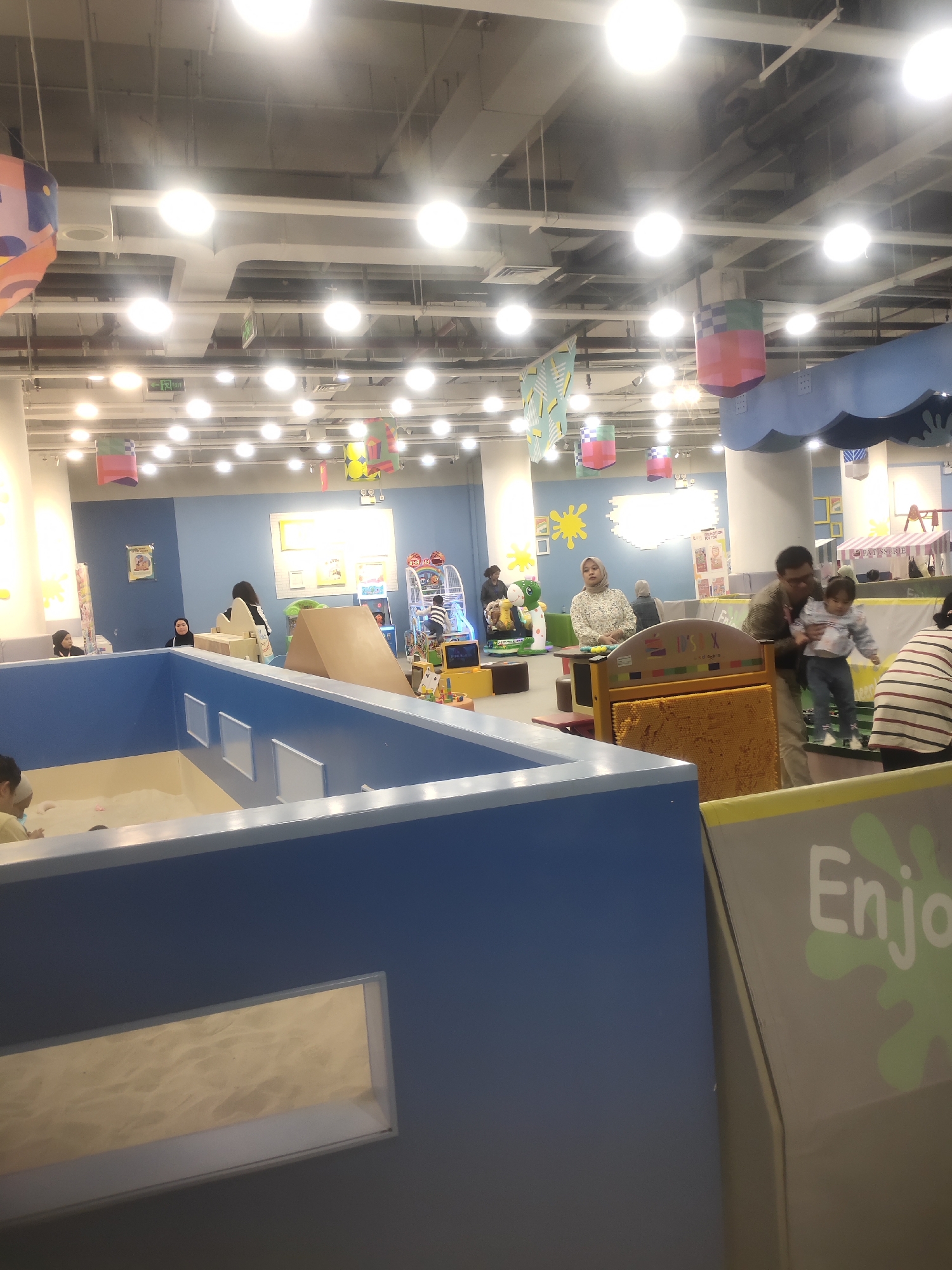 The image depicts the interior of a children's indoor playground, likely located in Bandung City, Indonesia. The environment is colorful and designed for play, with bright blue walls, numerous ceiling lights, and hanging decorative elements. In the foreground, a large blue structure with a sandpit area is visible. Several adults and children are present, engaged in various activities. One adult is holding a child, another is looking at a tablet, and others are standing or sitting around. Play equipment, such as a large pin art toy and a children's ride, are visible in the background. The overall scene suggests a lively and family-friendly atmosphere.