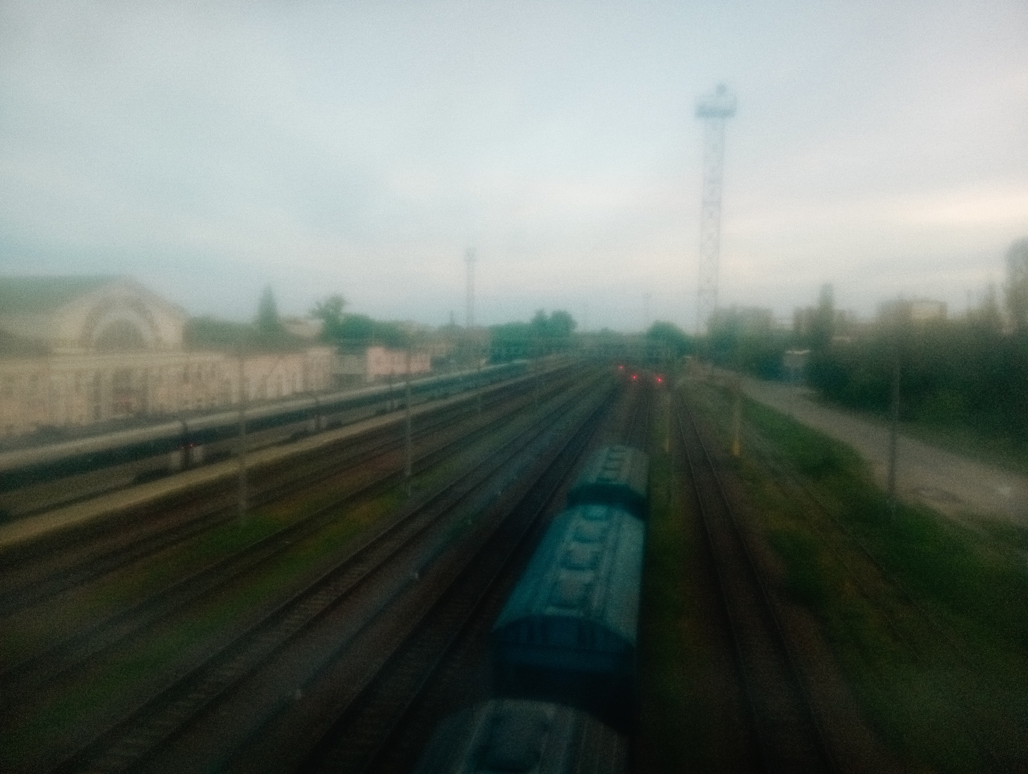 The image shows a view of the Poltava-Kyivska train station in Poltava, Ukraine, taken from an elevated position, likely a bridge or overpass. Multiple parallel railway tracks run through the scene, with a train partially visible in the foreground. A building with arched windows and a light-colored facade is visible to the left. A tall communications tower stands in the background amidst the hazy skyline. The sky is overcast. Vegetation is seen on the right side of the frame alongside a roadway.