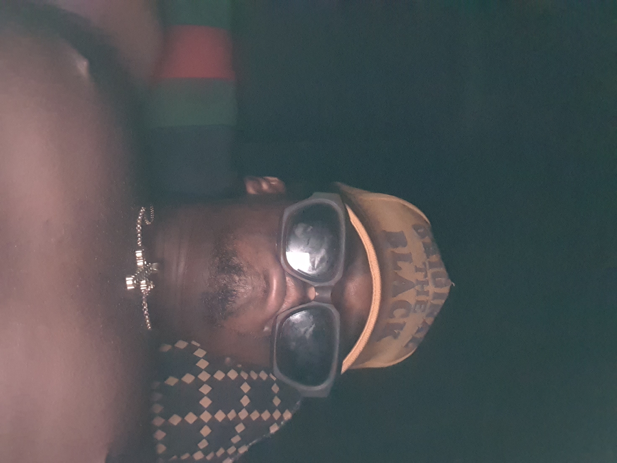 A man with brown skin, wearing sunglasses and a tan hat that reads "Protect the Black," is depicted. He is also wearing a silver chain with a cross. His shirt has a brown and black diamond pattern. Behind him is a blurry background of red, green, and black. The location in Lokoja, Nigeria is not discernible from the image.