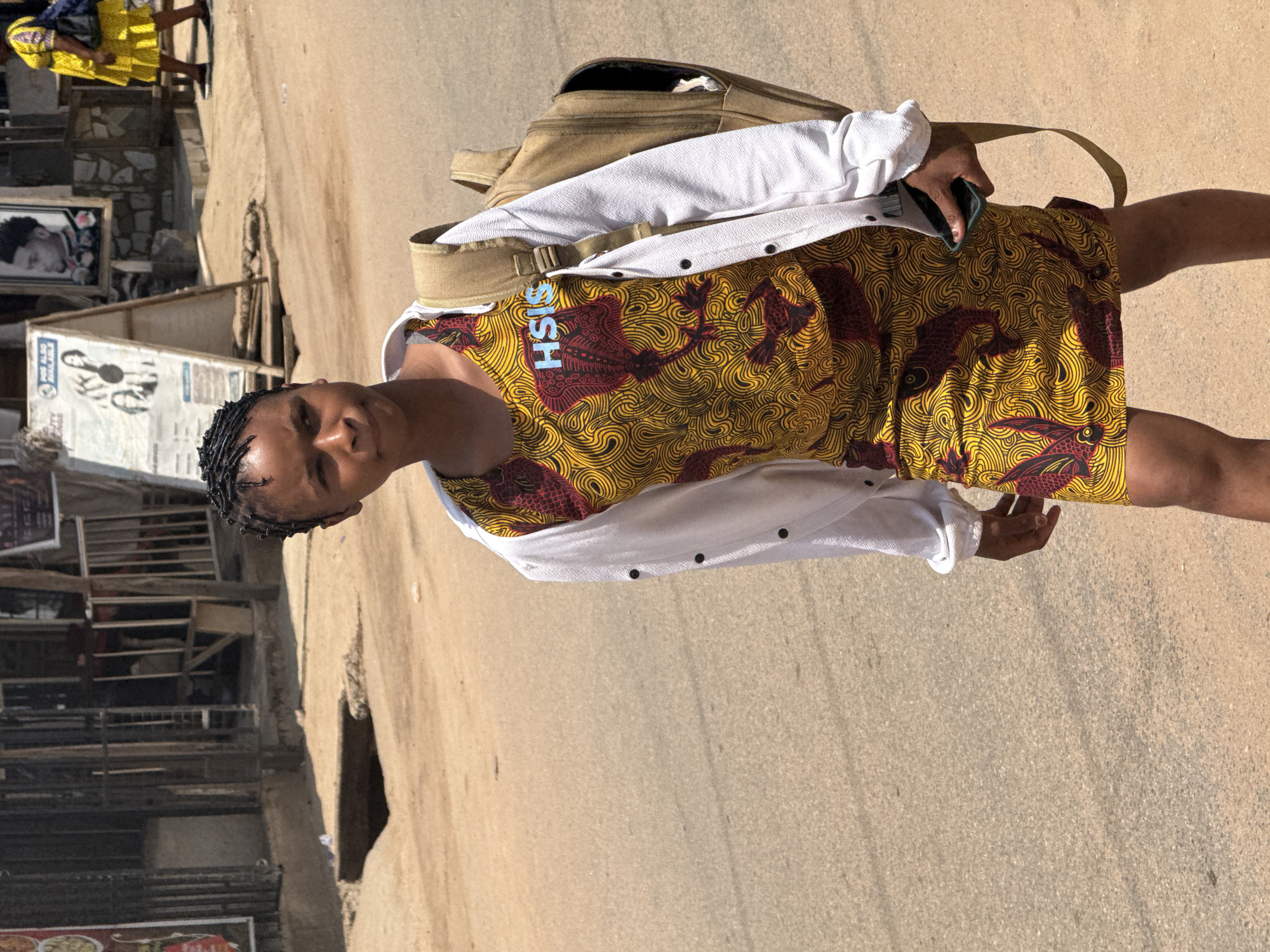 A young Black female stands on a dusty, paved road, possibly in Abuja, Nigeria. She is looking towards the right of the frame with a neutral expression. She has braided hair and is wearing a yellow and red patterned dress and a white open jacket. A light brown backpack is slung over her right shoulder. She holds a dark green cellphone in her right hand. In the background, there is a woman in a yellow dress, a stand with posters, and the facade of a building. The ground is covered in dirt and small pebbles, and there is a grated drain on the left side of the image. The lighting suggests it is a sunny day.