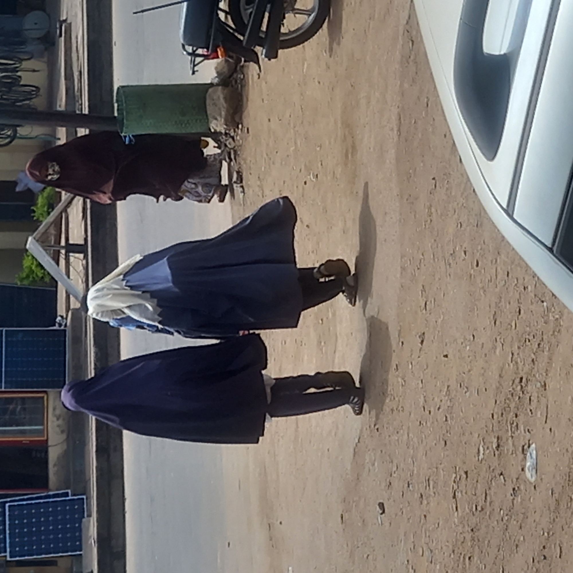 The scene depicts three individuals in Dadi Riba, Nigeria, walking on a dirt and gravel path beside a road. The person in the foreground is wearing a dark purple garment and black pants. The person in the middle is wearing a blue and white garment and black pants. The person in the background is wearing a brown garment and patterned pants. They are walking away from the camera, with a white car visible in the upper right corner and solar panels on the left side of the road. A motorbike and a green cylindrical object are visible further along the path.