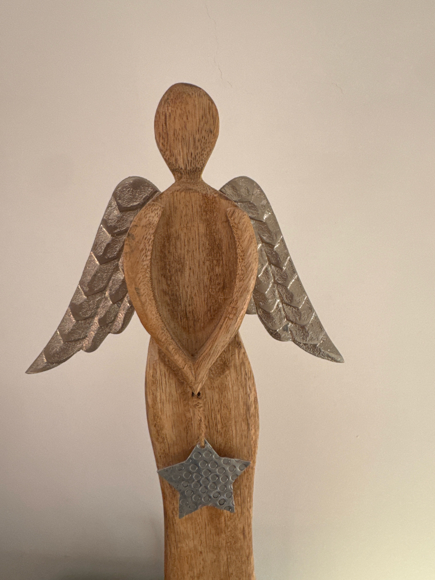 A wooden angel figurine with metal wings and a metal star hanging from its base is shown against a plain, light-colored background. The figurine is oriented facing away from the viewer, revealing its back. The background is a flat, neutral wall, devoid of any discernable features that would suggest a specific location within the city of Limassol, Cyprus. The lighting appears to be soft and diffused, casting minimal shadows. The image is a close-up shot, focusing solely on the angel figurine.