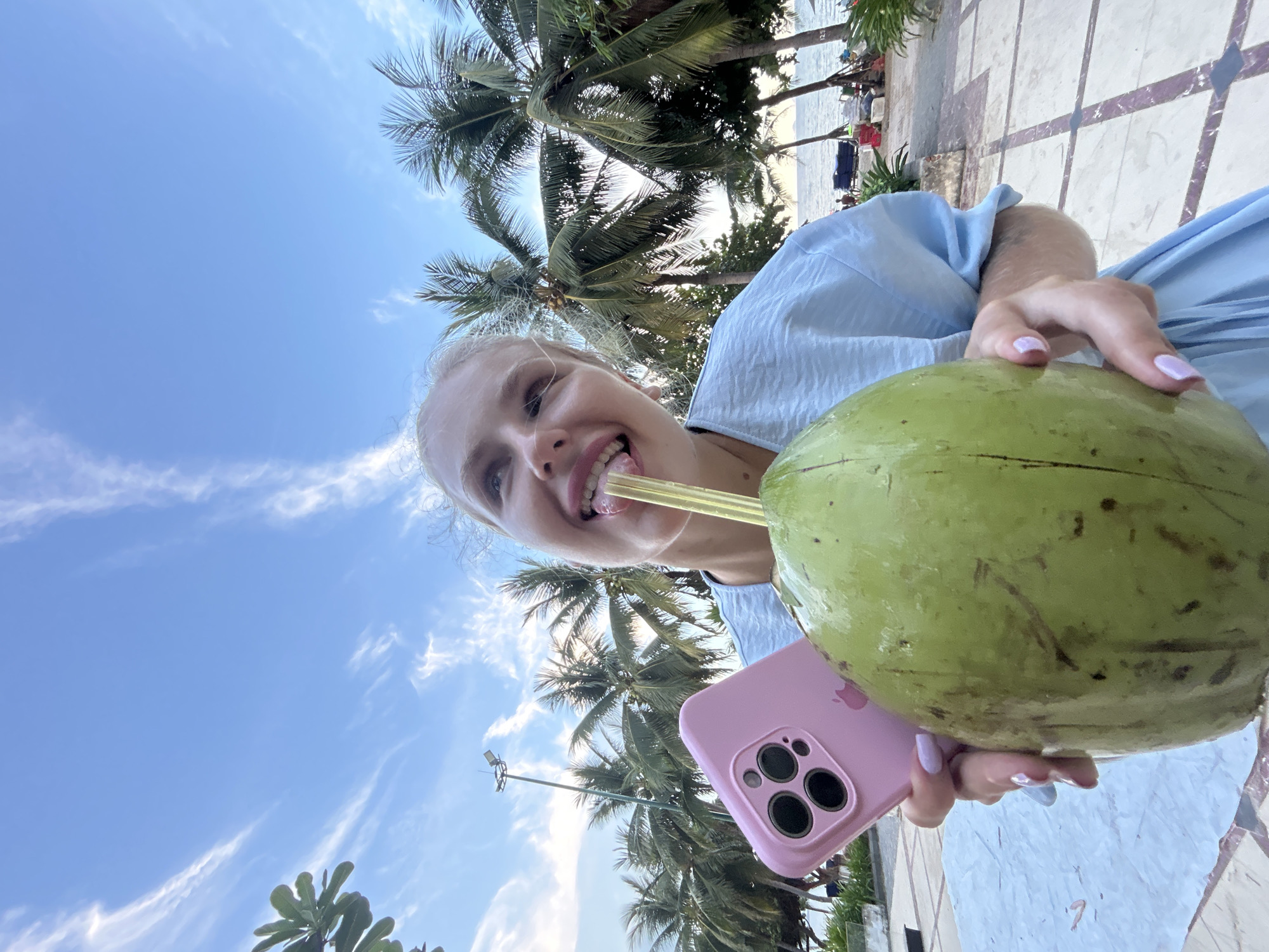 A woman with blonde hair, wearing a light blue dress, is shown from the chest up, holding a large green coconut with a straw. She is looking up and to the right with her tongue sticking out, touching the straw. In her left hand, she holds a pink iPhone case with three visible camera lenses. The background features a clear blue sky with wispy white clouds, numerous palm trees, and a glimpse of the ocean with a beach in the distance. The setting appears to be a beachside location, possibly in Nha Trang Ward, Vietnam, indicated by the presence of palm trees and the ocean.