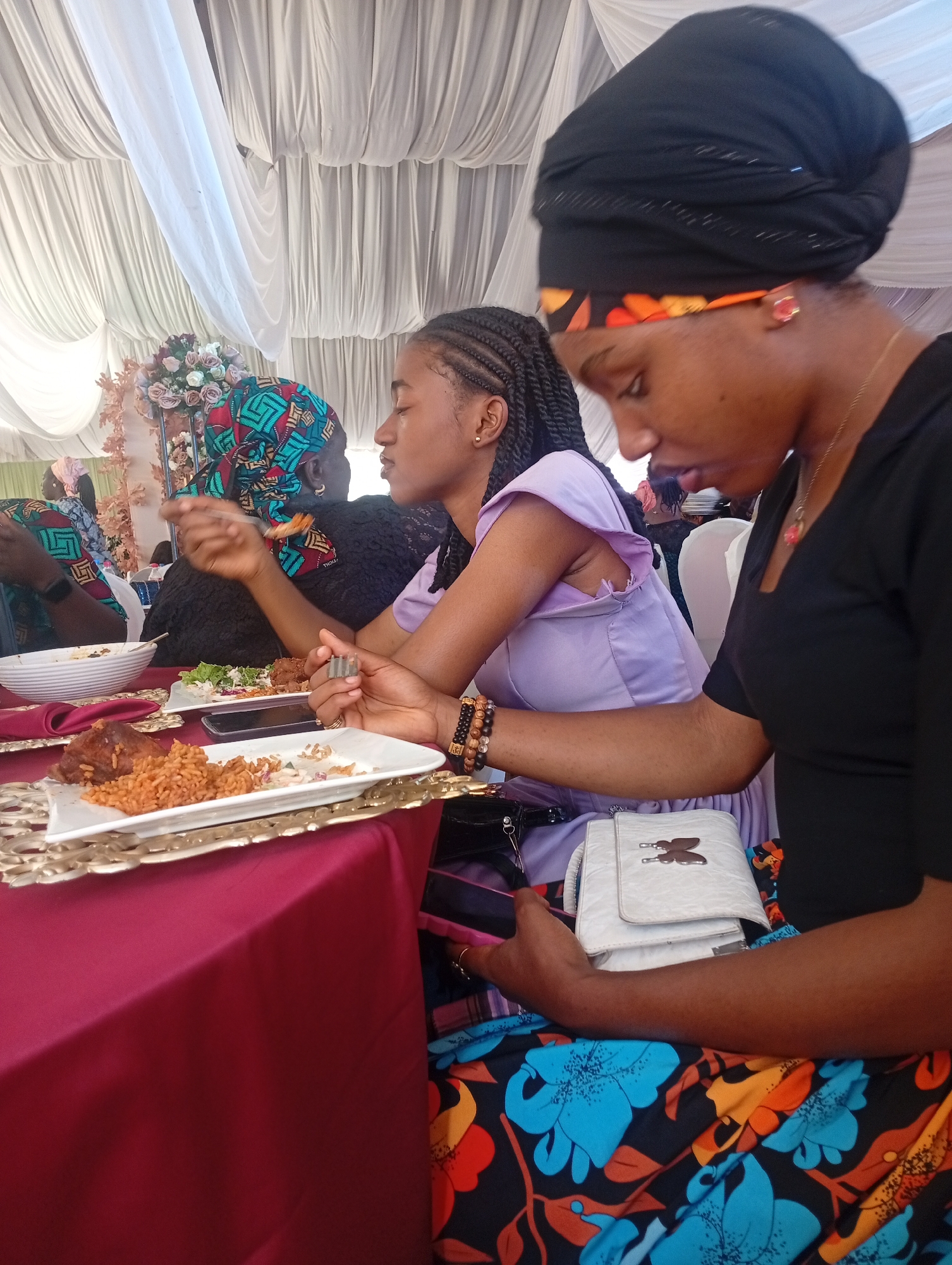 The image depicts a gathering of people in Barakin Lerawi, Nigeria, likely at a wedding or festive event, as suggested by the white draped ceiling. Three women are in the foreground, seated at a table covered with a maroon tablecloth. The woman on the right wears a black head covering with an orange band and a black dress, holding a white purse patterned with a butterfly design. The middle woman has braided hair and wears a lilac dress. The woman on the left wears a patterned head wrap and a black top. Plates of food, including what appears to be Jollof rice and fried chicken, are visible on the table. Other people can be seen in the background.