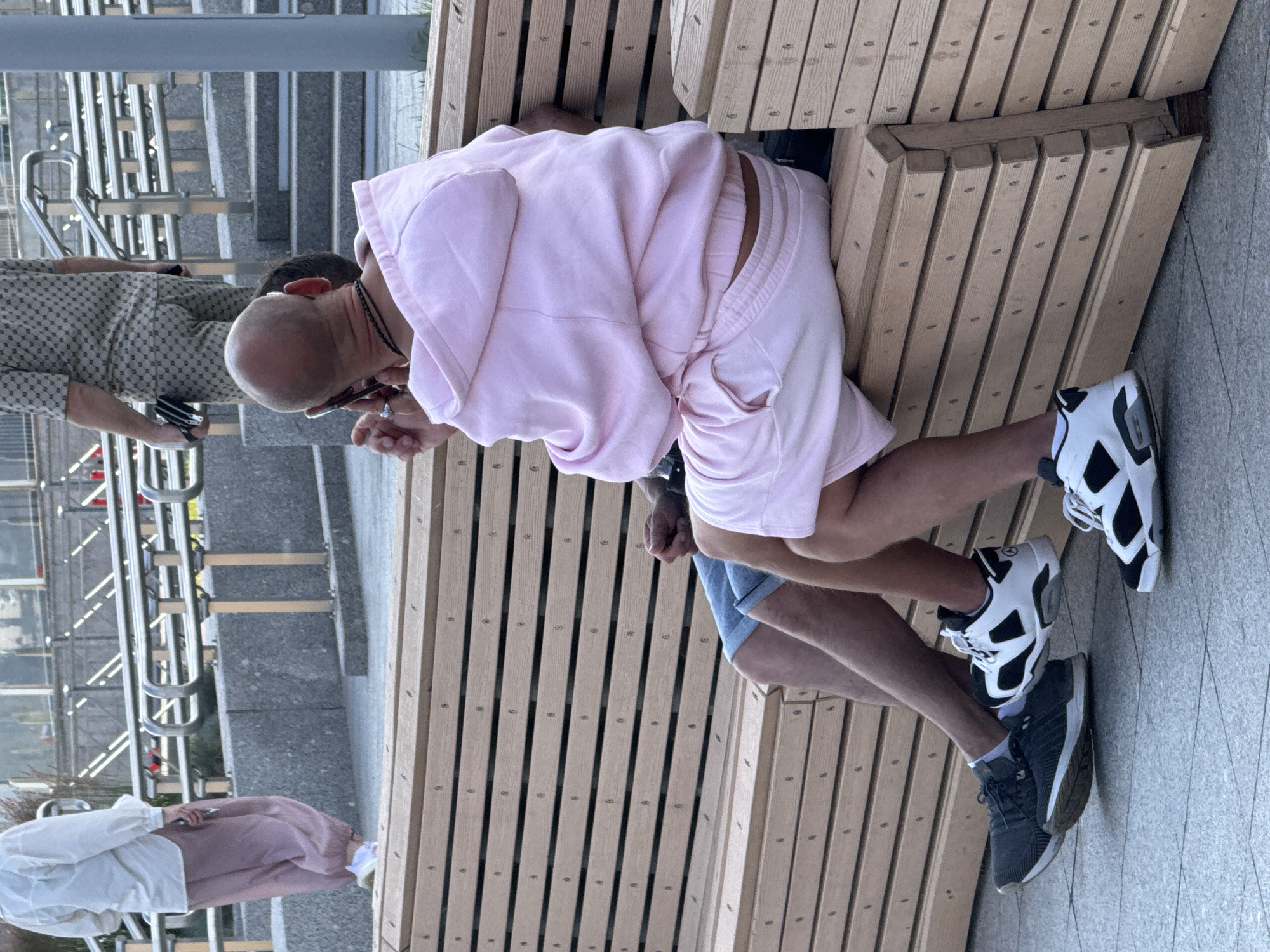 The image depicts two individuals sitting on a wooden bench in what appears to be an outdoor urban setting in Saint Petersburg, Russia. The foreground is dominated by the bench and the lower bodies of the two people. One person, viewed from the back, is wearing a pink hooded sweatshirt and pink shorts, and is on their phone. The other person, to their left and also partially obscured, is wearing denim shorts and athletic shoes. A third person is visible in the background walking up stairs, wearing a light-colored shirt and pink pants. The background shows modern architecture with metal railings and concrete structures, suggesting a public space like a park or plaza. The lighting suggests it is daytime.