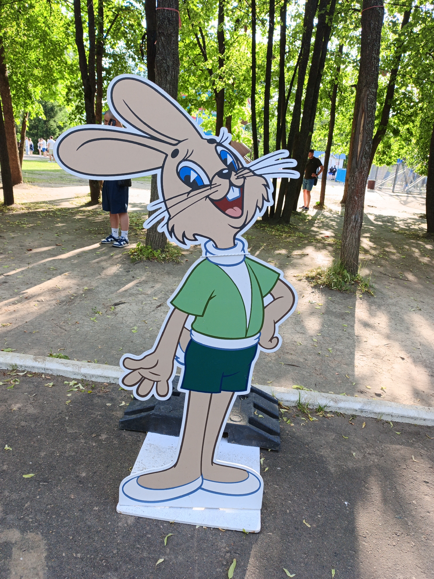 A cardboard cutout of a cartoon rabbit stands in a park-like setting. The rabbit is depicted wearing a green shirt with a white collar, dark shorts, and white shoes with blue trim. It has large ears, blue eyes, and a wide smile. The cutout is mounted on a white base and appears to be supported by black risers.
In the background, several trees with lush green foliage create a shaded area. Sunlight filters through the leaves, casting dappled shadows on the ground, suggesting it is daytime, likely late morning or afternoon. A few people can be seen in the distance, walking on a path. One person in the mid-ground is partially obscured by a tree and appears to be talking on a phone. Another person stands with their hands on their hips. Beyond them, a fence and some structures are visible. The ground is a mixture of asphalt and dirt, with fallen leaves scattered about. The overall scene evokes a casual, outdoor atmosphere, possibly at a fair, park, or recreational area. There is no visible text in the image.