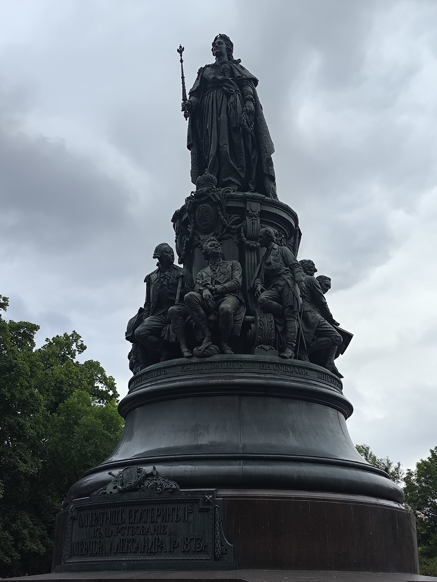 This is a medium-shot, low-angle, outdoor photograph of a large bronze monument. The monument is dark and appears to be made of metal. It is topped by a statue of a woman in a long, draped gown, wearing a crown and holding a scepter. Below the central statue, several smaller bronze figures of men are seated or standing around the base of the monument. Some of these figures are depicted in military attire. The monument is very detailed, with ornate carvings and inscriptions visible on its various levels.
The monument is set against a cloudy, overcast sky. To the left and right of the monument, lush green trees are visible, suggesting an outdoor park or public square setting. The overall mood of the image is somber, likely due to the dark monument and the cloudy weather.
Text is visible on the base of the monument. The most prominent inscription, in Cyrillic script, appears to read: "ИМПЕРАТРИЦЕ ЕКАТЕРИНЫ II ВЪ ЦАРСТВОВАНИЕ ИМПЕРАТОРА, АЛЕКСАНДРА II 1873." This translates to "To Empress Catherine II during the reign of Emperor Alexander II 1873." Other inscriptions with names like "КН. СУВОРОВЬ" are also partially visible. The style of the monument and the inscriptions suggest it is a historical monument, possibly commemorating Empress Catherine the Great and Emperor Alexander II.