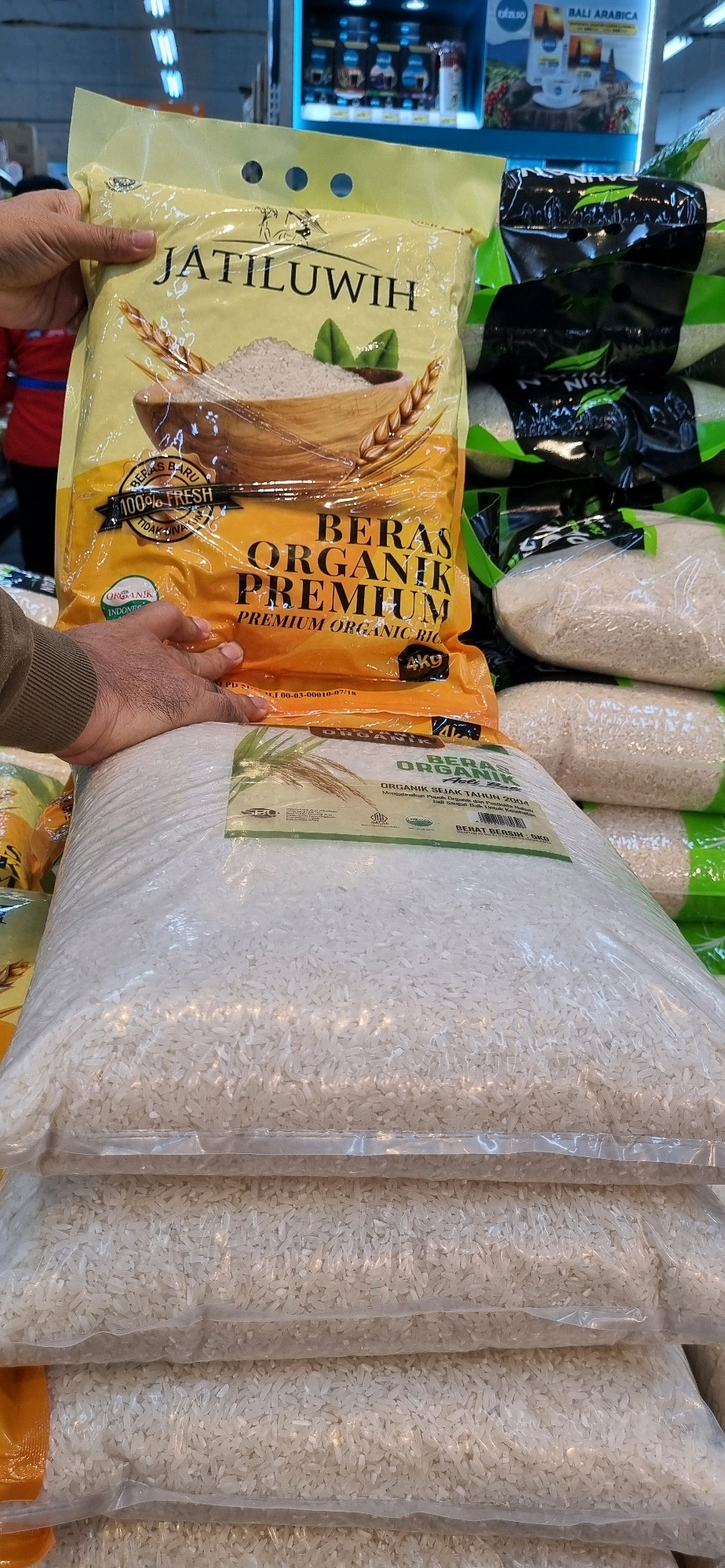 A person's hand is holding a large, yellow bag of "JATILUWIH BERAS ORGANIK PREMIUM" (Jatiluih Premium Organic Rice). The bag displays images of rice grains, wheat stalks, and leaves, with text in Indonesian indicating it is 100% fresh and organic. Next to the yellow bag, there are stacks of large, clear plastic bags filled with white rice, also labeled with "ORGANIK" (Organic). The background shows shelves with products, including what appear to be coffee packages and a sign for "BALI ARABICA." The scene is likely within a supermarket or grocery store in Denpasar, Indonesia, given the mention of "Jatiluih" which is a famous rice-terraced area in Bali, and the context of a retail environment with consumer goods. The lighting suggests an indoor setting, possibly with fluorescent or display lighting typical of a store. The image captures a consumer product display of organic rice.
