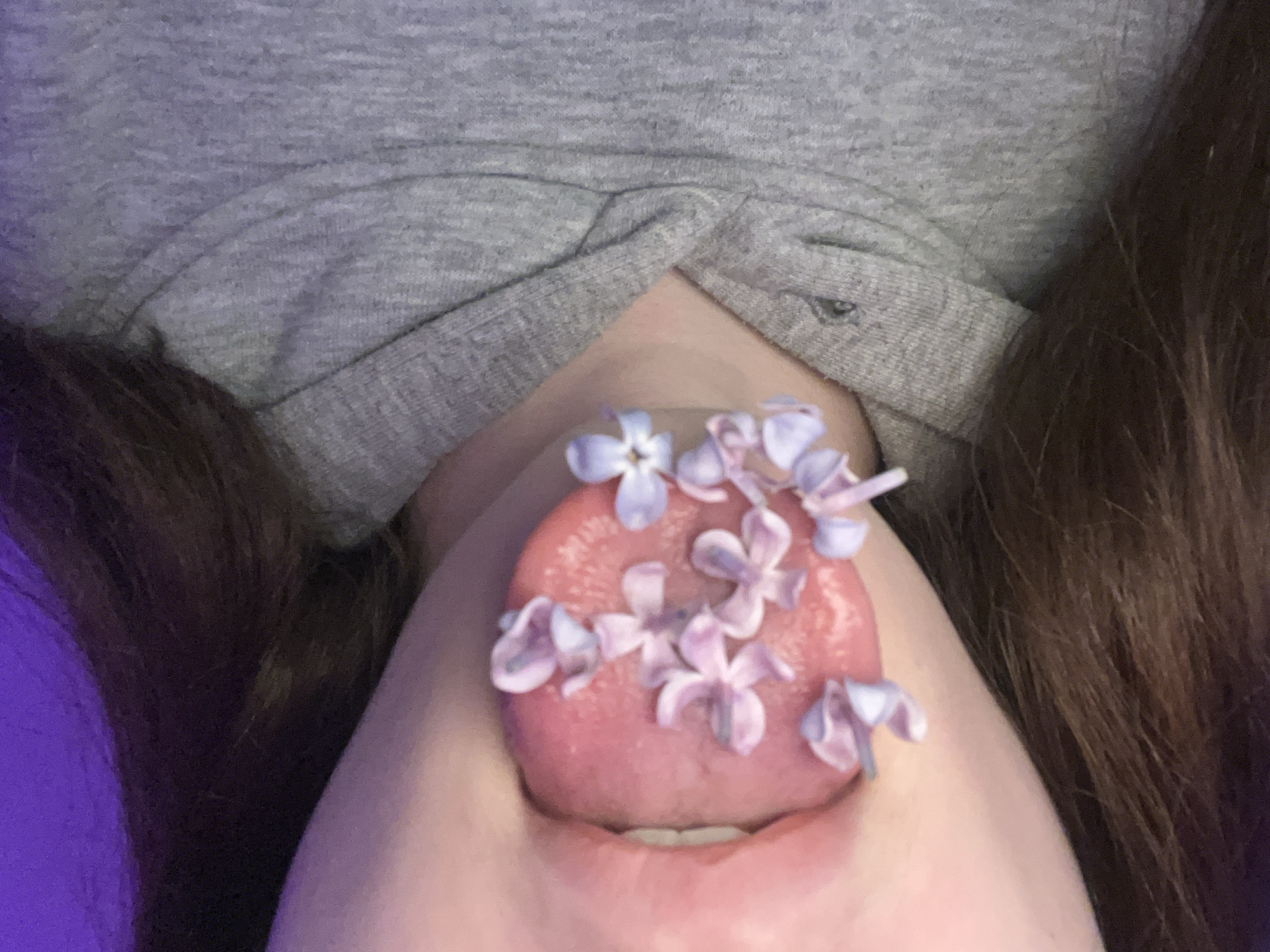 The image shows a person's open mouth with their tongue sticking out, covered in small purple and white flowers. The person is wearing a grey shirt, and brown hair is visible on the sides of the frame. The background is blurry. There is no indication of location in Saint Petersburg, Russia.