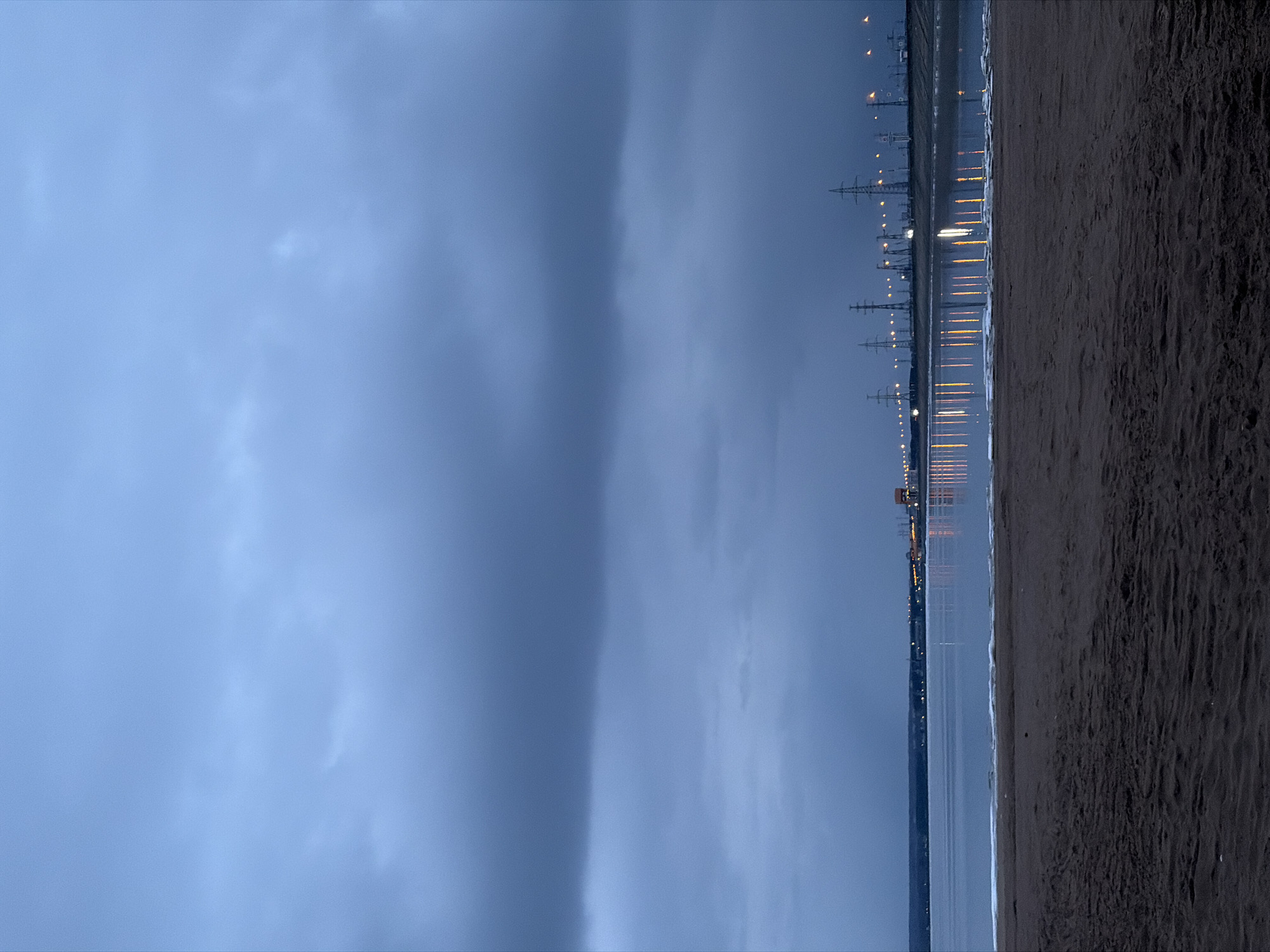 The image depicts a beach scene at twilight in Noviy, Russia. The sky is overcast and blue-grey. To the right of the image, there is a dark, sandy beach. A body of water separates the beach from a distant shoreline, where lights from buildings and transmission towers are visible. Reflections of the lights are seen on the water's surface.