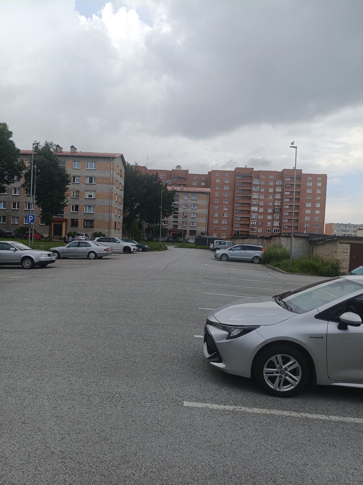 The image shows an outdoor parking lot in Narva, Estonia, with several cars parked. In the background, there are residential buildings with multiple stories. The sky is overcast with clouds. A silver car is parked in the foreground, partially obscuring the view of other vehicles and the parking lot markings. Other cars visible include silver sedans and a white SUV. Trees are present on the left side of the image, and a brick wall is visible behind some of the parked cars. A street light is also present in the scene.