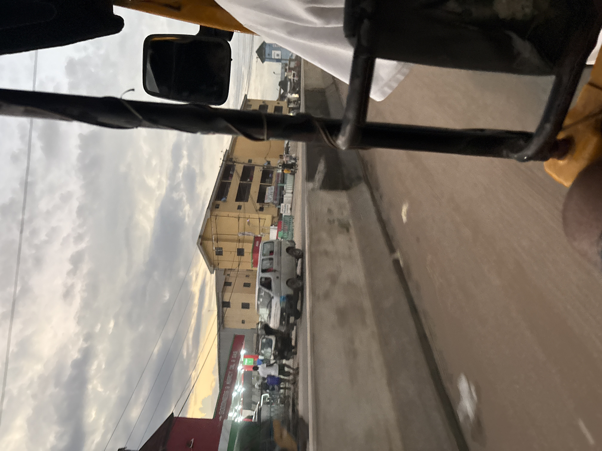 The image depicts a view from within a rickshaw in the city of Onitsha, Nigeria. The scene captures a street view with buildings, vehicles, and people. In the foreground, part of the rickshaw, including its black frame and yellow body, is visible. A white cloth is draped over the seat in the background. The street is lined with a concrete barrier. On the other side of the barrier, a red building with green accents is present, possibly a shop. Further down the street, a yellow, multi-story building stands. Several vehicles, including a white van and other smaller vehicles, are on the road. A few individuals are walking and standing near the shops. The sky above is overcast with clouds. The overall impression is of a bustling street scene in a Nigerian city.