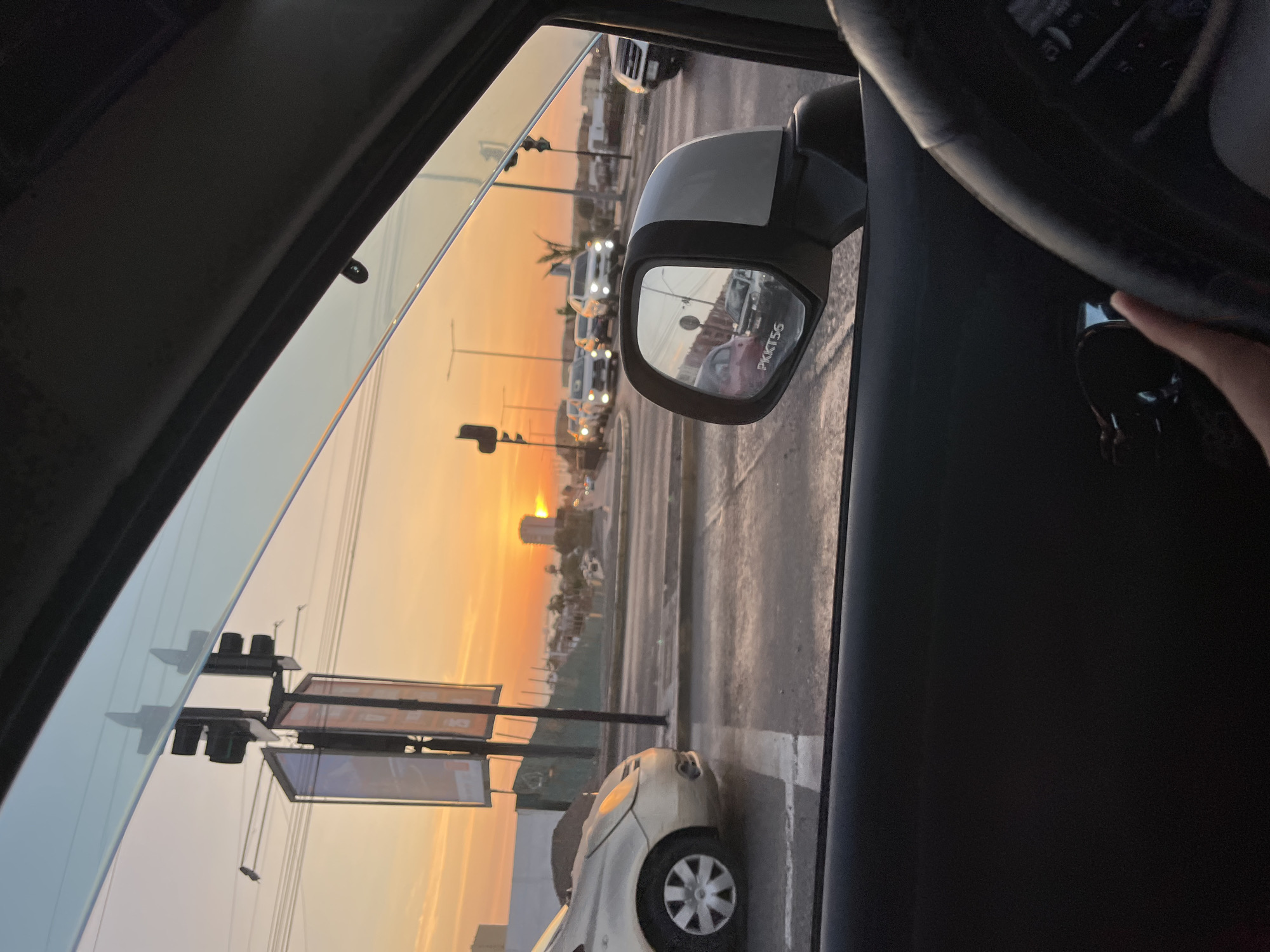 The photo shows a view from inside a car at sunset, in Arica, Chile. The sun is setting over a cityscape with buildings, traffic lights, and palm trees. A line of cars is visible on the road, reflected in the car's side mirror. The car's steering wheel and dashboard are partially visible in the foreground. The light is warm and orange, typical of a sunset.