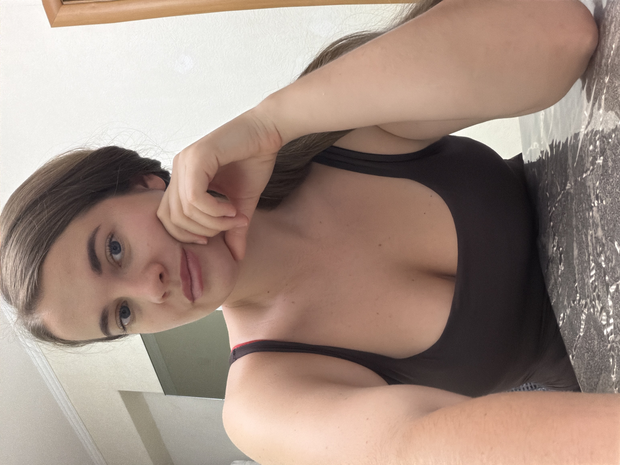 A fair-skinned young woman is posing for a selfie in an indoor setting. She has medium-length brown hair and blue eyes. She is wearing a dark-colored tank top with a low neckline. She is holding her hand to her face. The background is blurry and contains a light-colored wall and a piece of light-colored furniture at the top. A grey surface with patterns is on the right. The location cannot be identified from the image.