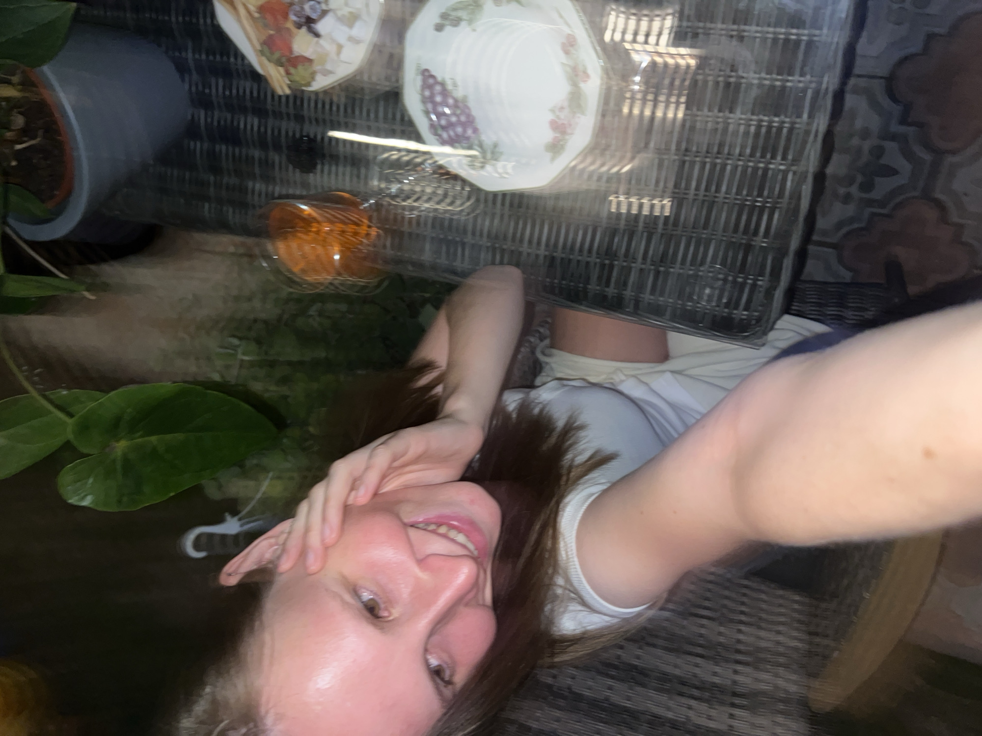 The media file is a selfie taken by a young woman lying down. She is smiling and looking at the camera, with her hand resting on her head. She is wearing a white top. In the background, there is a wicker table with a glass top, on which are two decorative plates with fruit designs, a glass of amber liquid, and what appears to be a small cage. To the left of the table is a potted plant with large green leaves. The scene appears to be outdoors, possibly in a garden or patio area, with patterned tiles visible on the ground in the background. The lighting suggests it might be evening or nighttime. There is no visible text in the image.