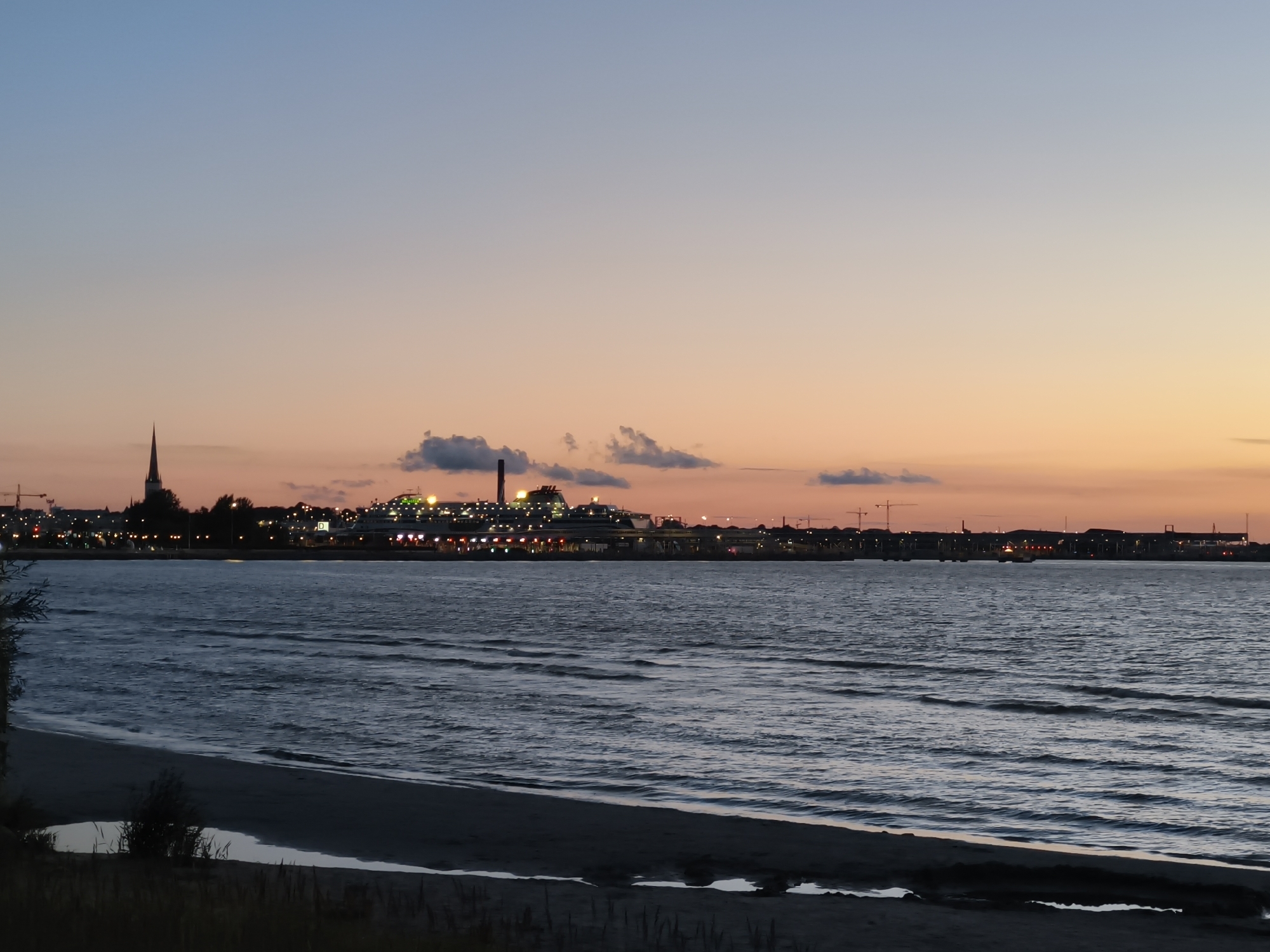The image depicts a twilight view of the Tallinn Bay waterfront, with the city skyline visible in the distance. The sky transitions from a pale blue at the top to shades of pink and orange near the horizon, indicating sunset or sunrise. In the foreground, a calm body of water, likely the bay, is shown with gentle ripples. A dark, sandy beach is visible along the bottom edge of the frame, with some sparse vegetation and a small puddle of water.
The city skyline features several prominent structures. To the left, a church spire with a pointed roof stands out against the darkening sky. Further to the right, a large ferry or cruise ship is illuminated with numerous lights, docked at the port. The silhouette of buildings and industrial structures, including cranes, are visible along the coastline, suggesting a busy port area. The overall scene conveys a peaceful, urban waterfront setting at dusk. There are no people or animals clearly visible in the image.