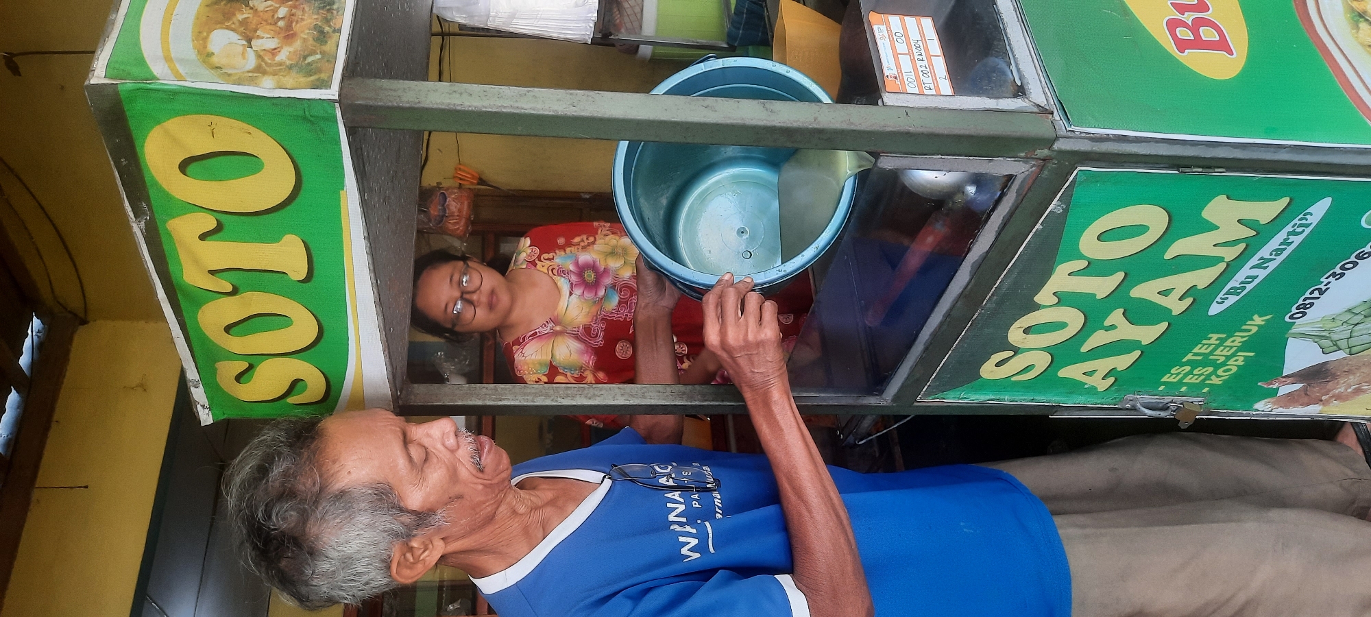 In the city of Selopuro, Indonesia, a man with gray hair and a mustache, wearing a blue t-shirt and tan pants, is pouring liquid from a light blue bucket into a food stall. Behind him, a woman wearing glasses and a colorful floral-patterned shirt is standing. The food stall has green signage that reads "SOTO AYAM" in yellow lettering, and also lists "ES TEH," "ES JERUK," and "KOPI." A small label is affixed to the stall with text including "DD11," "00," and "PT 002 RW.004 2 1". The stall appears to be outdoors, with a yellow wall and ceiling visible in the background.