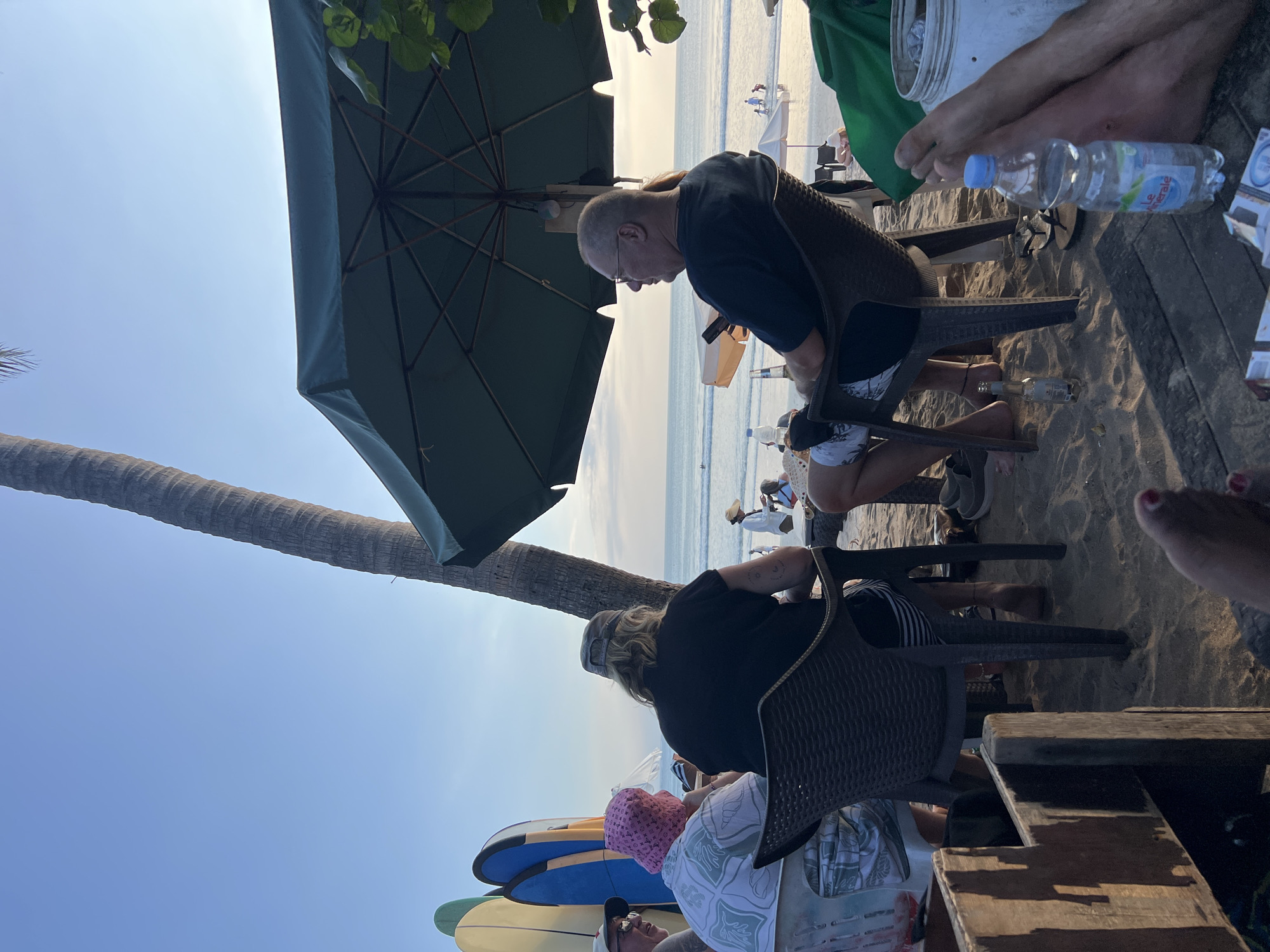 This image captures a group of people at a beachside establishment in Kuta, Indonesia, during what appears to be late afternoon or early evening. The scene is set on a sandy beach with the ocean visible in the background, featuring gentle waves and a few people in the water.
In the foreground, several individuals are seated at tables, enjoying beverages and the ocean view. One man, wearing glasses and a dark t-shirt, is seated facing right, looking out towards the sea. Next to him, another person with blonde hair and a cap is seated with their back to the camera. To the left, two individuals are visible, one wearing a pink patterned hat and a white shirt with a green design, and another person partially obscured. Surfboards are propped up behind them, suggesting a surf-centric location.
Underneath a large, dark green umbrella, providing shade, the tables are set on the sand. A bottle of water and what appears to be an empty beer bottle are visible on a wooden table in the lower right portion of the frame, along with cigarette packs. The overall atmosphere suggests a relaxed setting, likely a beach bar or cafe. The sky is clear with a hint of sunset colors starting to appear. A prominent palm tree trunk is visible on the right side of the image, adding to the tropical ambiance of Kuta.