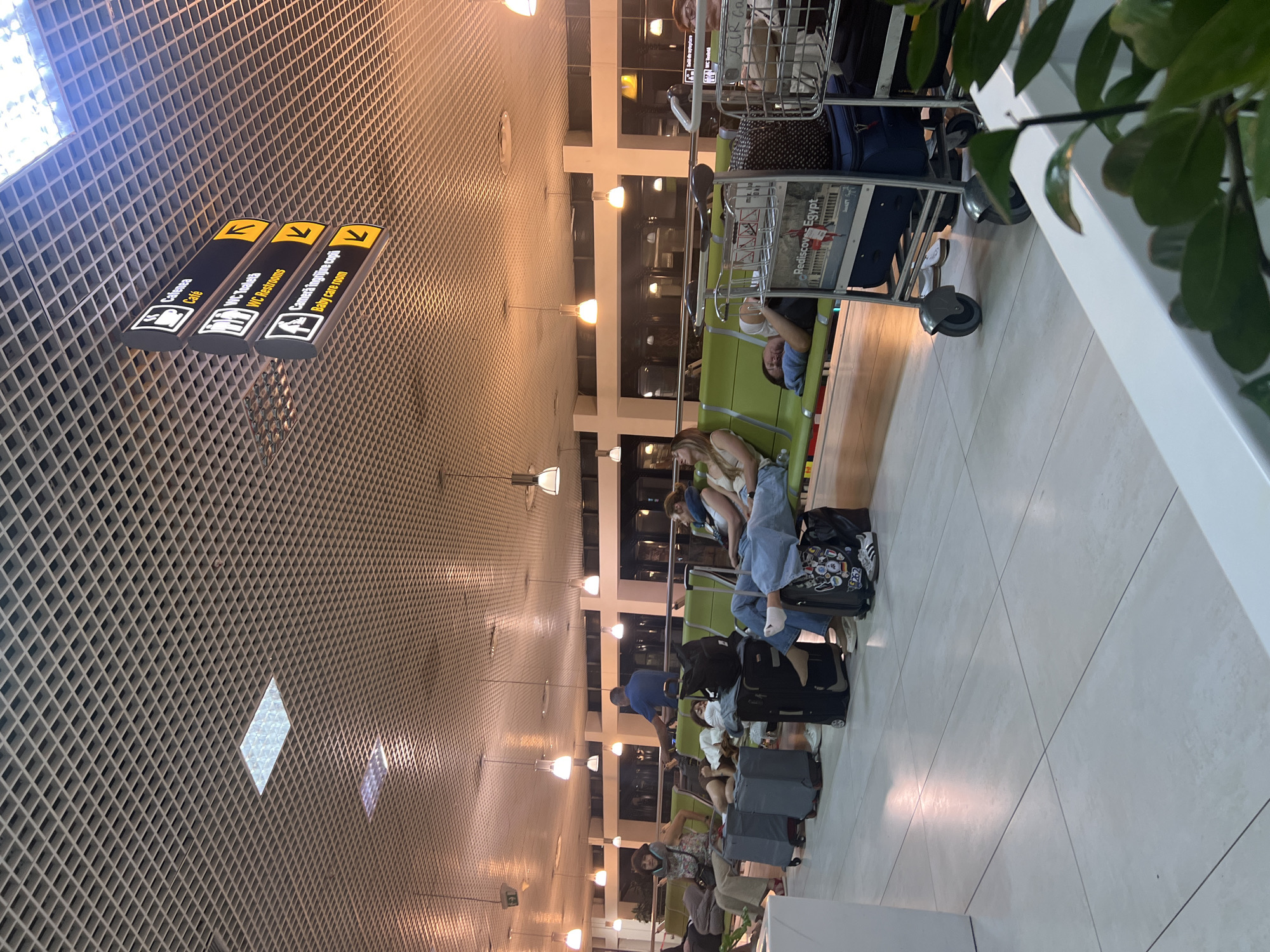 The image depicts an airport waiting area, likely within Chisinau International Airport in Chisinau, Moldova, based on the presence of directional signs in Romanian and English. Several travelers are seen resting on green airport seating. One person is lying down with their head on a pillow, while others are seated, some with their heads tilted back or resting on neck pillows. Luggage, including suitcases and a shopping cart filled with bags, is present near the seated passengers. Overhead, a grid-like ceiling with embedded lighting fixtures and signs is visible. One sign indicates directions for "Café," "WC Restrooms," and "Baby care room." The floor is tiled with large, light-colored tiles. The overall atmosphere suggests a waiting period, possibly due to a flight delay or early arrival at the airport.