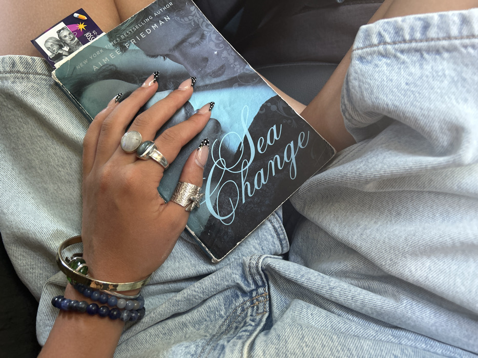A person sits wearing light-wash denim shorts, holding a book titled "Sea Change" by Aimee Friedman. The hand on the book is adorned with multiple rings, including one with a large moonstone. The nails are long with a clear base and black polka dot tips. A silver bangle with a green stone and two beaded bracelets decorate the wrist. A small card with a picture of an elderly couple is tucked into the book's pages.