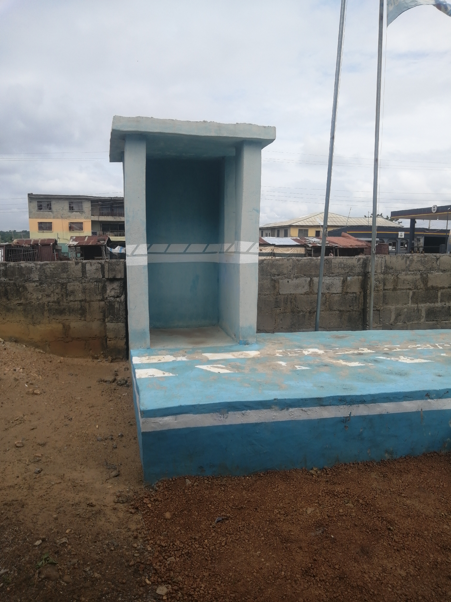 A blue and white concrete structure, resembling a shelter or seating area, stands on a dirt and gravel ground. The structure has a flat roof supported by two pillars. The interior of the structure is painted light blue, with a white horizontal stripe pattern on the back wall. The exterior of the structure, including a wide base, is painted in alternating bands of light blue and white. Behind the structure, a grey brick wall runs horizontally, and further in the background, there are buildings, including a multi-story structure with balconies and a gas station. Two flagpoles stand to the right of the structure, with a partially visible flag at the top. The sky is overcast.