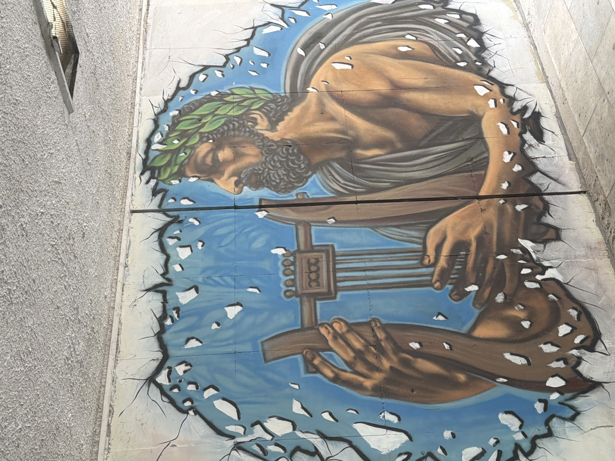 A mural on a wall in Plovdiv, Bulgaria, depicts a person with a beard and a laurel wreath on their head, dressed in a gray toga, playing a lyre. The mural is designed to look like it is bursting through the wall, with cracks and broken pieces around the edges. The background is blue, possibly representing the sky. The mural appears to be painted on rectangular tiles or sections of the wall. Part of a gray, textured building is visible on the left side of the image. The entire scene is viewed at an angle.
