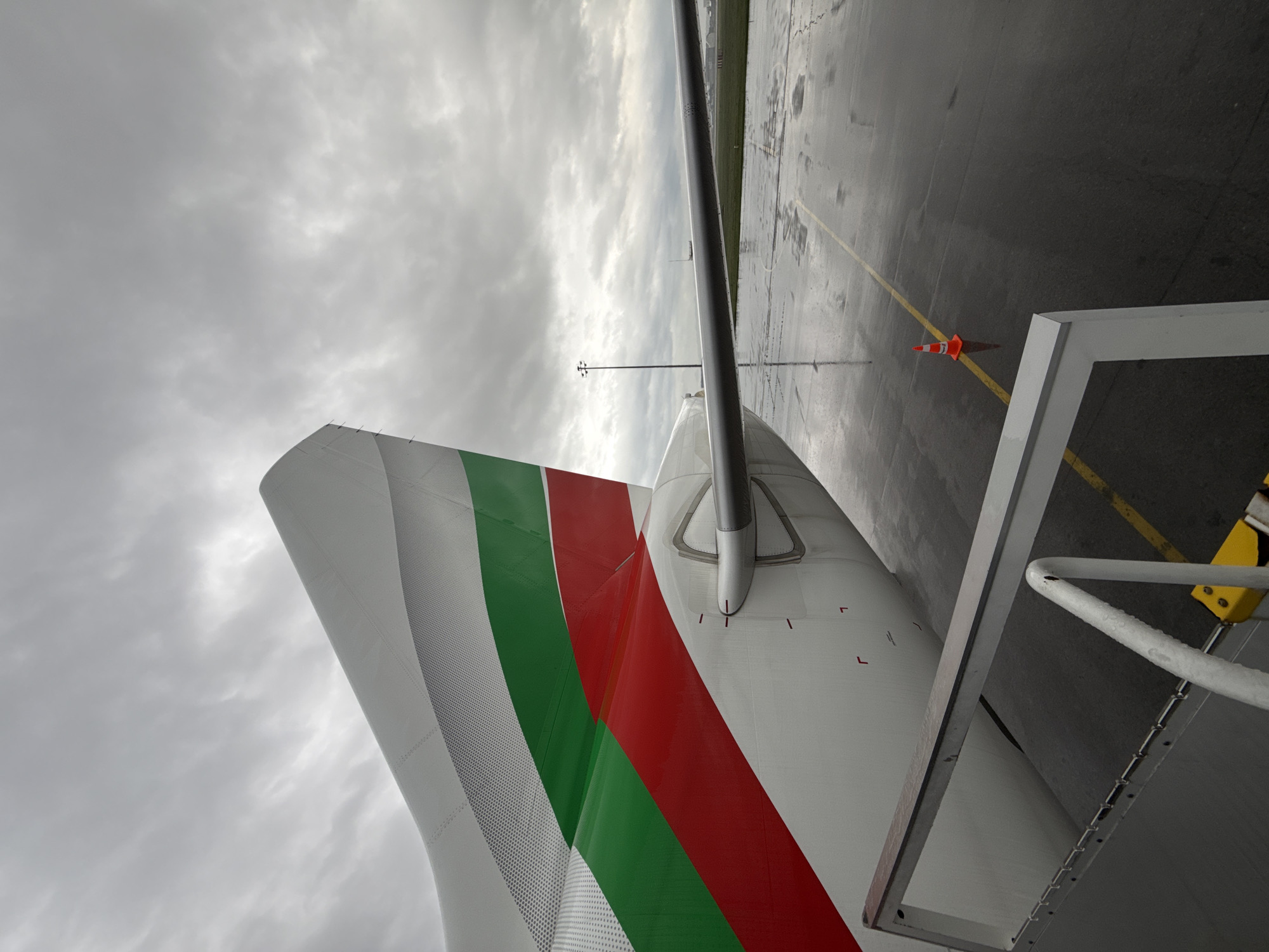 The image shows the tail of an aircraft, painted with white, green, and red stripes, colors which match the flag of Bulgaria. The aircraft is parked on a wet tarmac, with visible painted lines and an orange cone nearby. A metal staircase or platform is positioned adjacent to the aircraft's tail. The sky is overcast with heavy cloud cover. Given the Bulgarian flag colors on the tail of the aircraft, it is highly probable the location is at Sofia Airport in Sofia, Bulgaria.