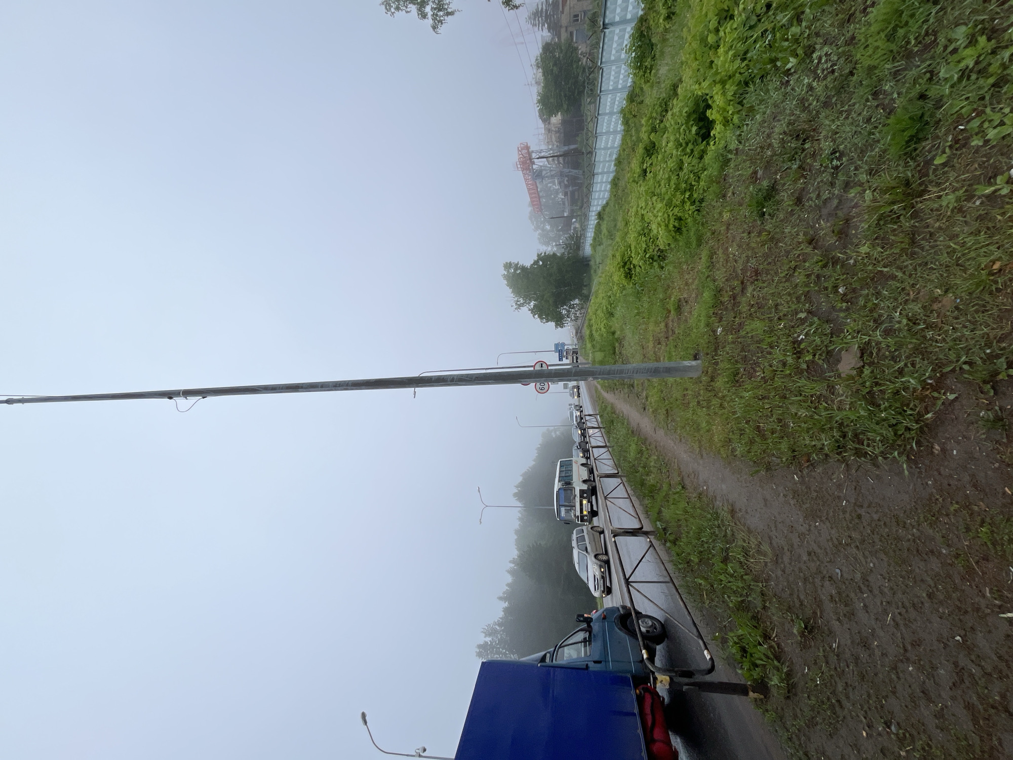 This image depicts an outdoor scene in Chaikovsky, Russia, on a heavily foggy or misty day, suggesting a cool and damp atmosphere. The ground, including a dirt path and grassy areas, appears wet and muddy.
In the foreground, a tall utility pole runs horizontally across the top of the image (when viewed upright). Below it, a line of vehicles, including a blue truck partially visible on the far left, several cars, and a white bus, are stationary or moving slowly, indicating traffic or a queue. A metal barrier or fence separates the road from a narrow dirt path and a steep, grassy embankment on the right side of the road. A circular red-bordered speed limit sign, clearly displaying "60," is visible near the road.
The midground and background are significantly obscured by the dense fog, reducing visibility. Through the haze, outlines of distant industrial structures, possibly cranes or large buildings, can be faintly discerned. A patterned green and white barrier or wall runs along the top of the embankment in the distance, adjacent to more trees and foliage. The overall scene conveys a quiet, muted environment due to the pervasive fog.