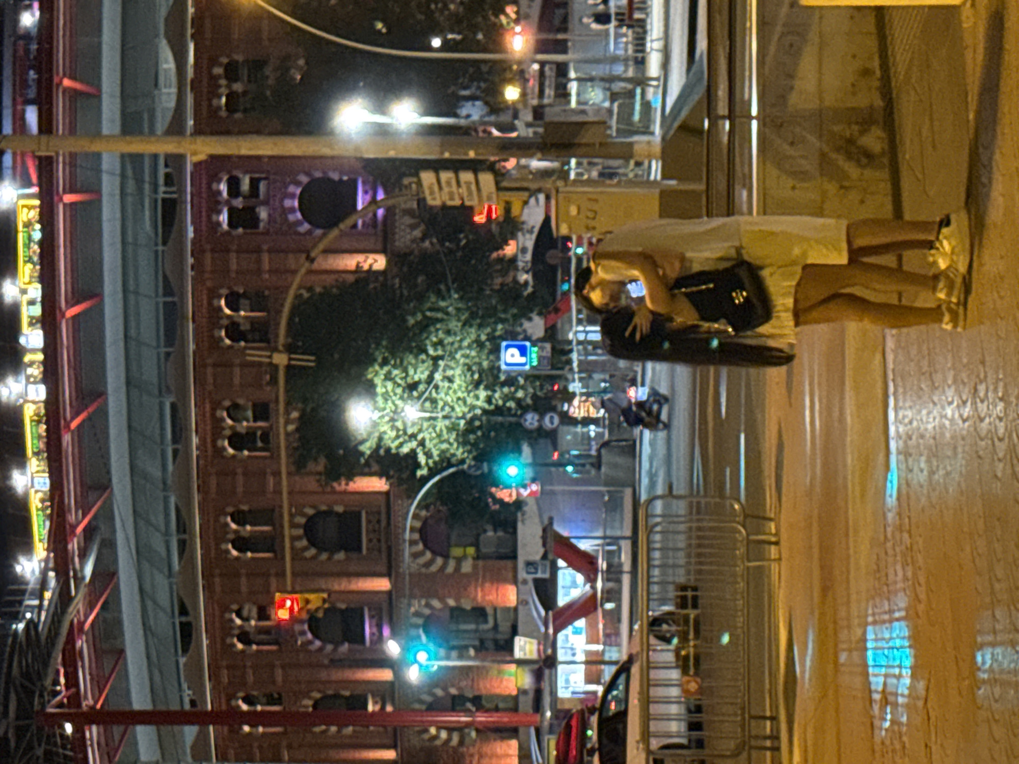 The image depicts an urban street scene at night in Barcelona, Spain, likely near the Las Arenas de Barcelona building, given the architectural style of the brick structure in the background. A couple is in the foreground, embracing and kissing. The woman, on the left, has long dark hair and is wearing a light-colored skirt and a dark top, carrying a black purse. The man, on the right, is wearing a light-colored t-shirt and white shorts. They are standing on a wet pavement, reflecting the ambient light from streetlights and building illuminations.
In the mid-ground, a white car is parked behind a metal barrier. A scooter with a rider is moving away from the camera towards the right. The background features a large, circular, brick building with arched windows, illuminated by warm lights. Above this building, a structure with red beams and a blue roof is visible, likely part of the Las Arenas complex. Traffic lights show red and green signals. Streetlights, signs, and trees add to the urban night atmosphere. The ground is wet, suggesting recent rain or street cleaning.