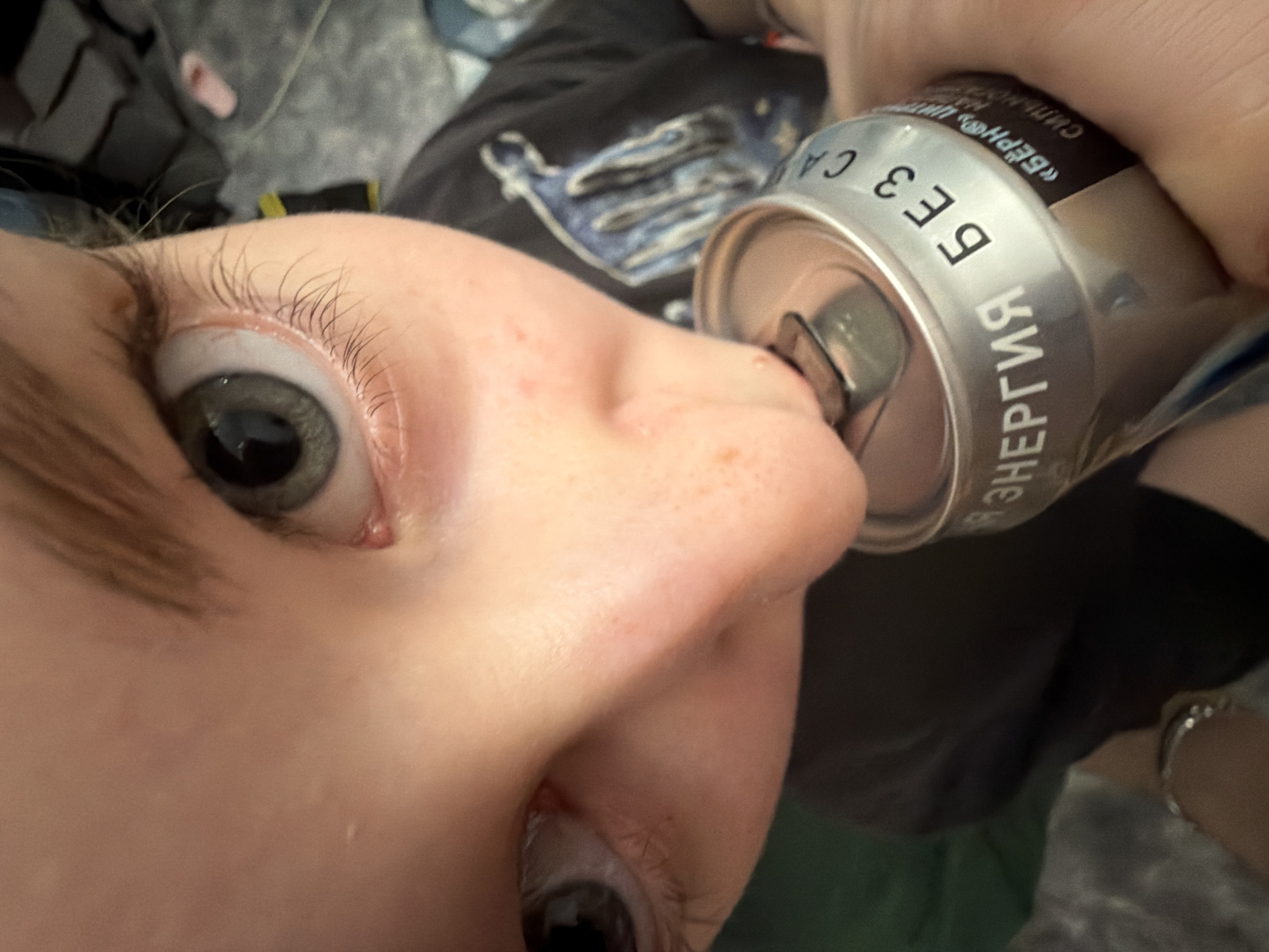 A close-up, low-angle shot depicts a person drinking an energy drink. The person's face is prominent, with a focus on the left eye and nose. The energy drink can is silver and labeled in Russian. The text on the can includes the words "БЕЗ СА" and "ЭНЕРГИЯ." The background is blurred, showing a dark-colored T-shirt. The environment is unclear; the location of Perm, Russia, cannot be identified from the image.