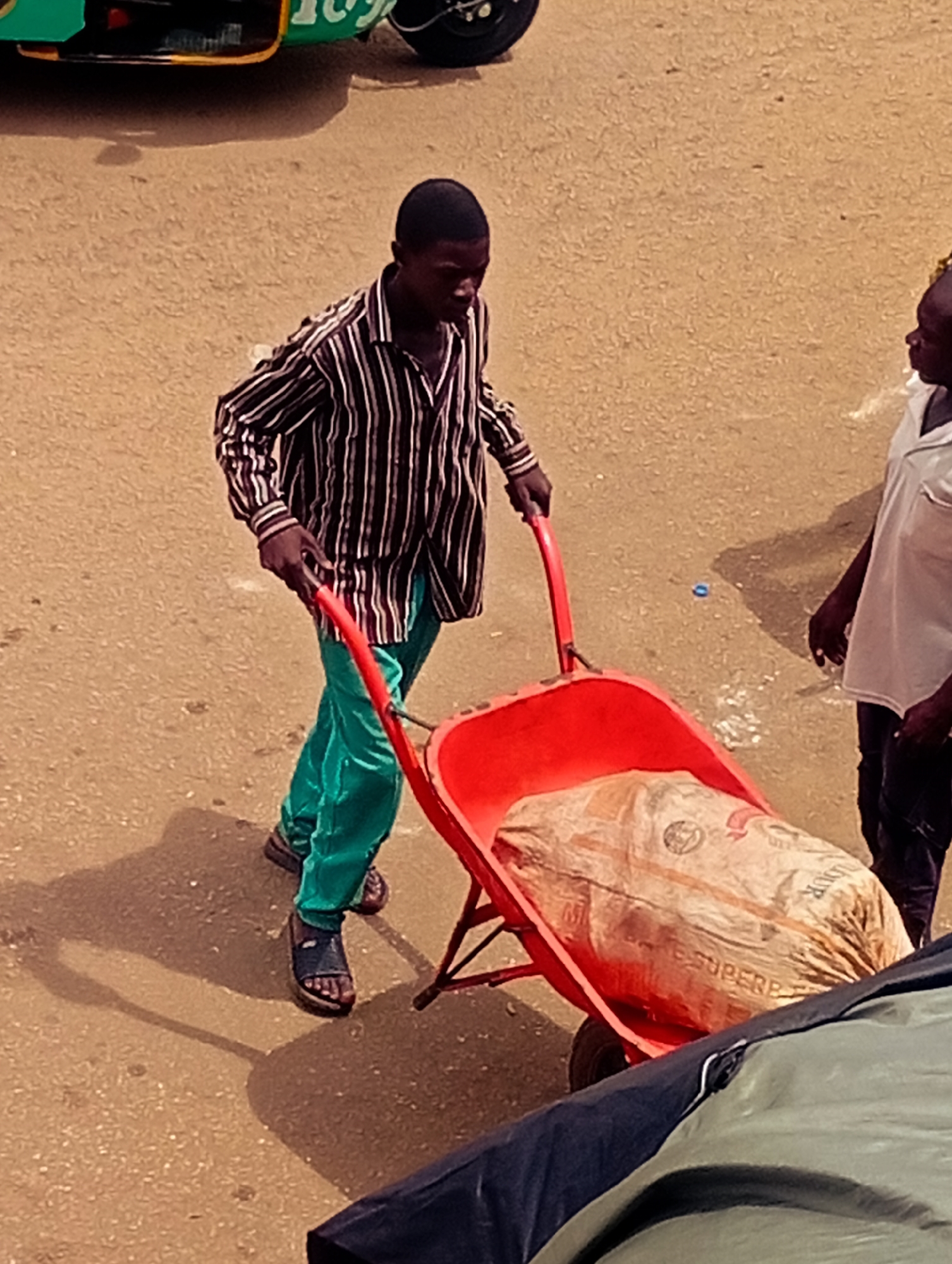 In Bukuru, Nigeria, a young Black man wearing a black and white striped shirt and bright teal pants pushes a red wheelbarrow filled with a large bag of cement. He is wearing dark sandals. To his right, another person, also Black and wearing a white shirt and dark pants, stands watching. The ground is a dusty, sandy surface. A green and black vehicle is partially visible in the upper left corner. The scene appears to be outdoors in daylight, possibly a market or construction area. The overall mood seems to be one of labor and daily life. Text is visible on the cement bag, including "SUPERB".