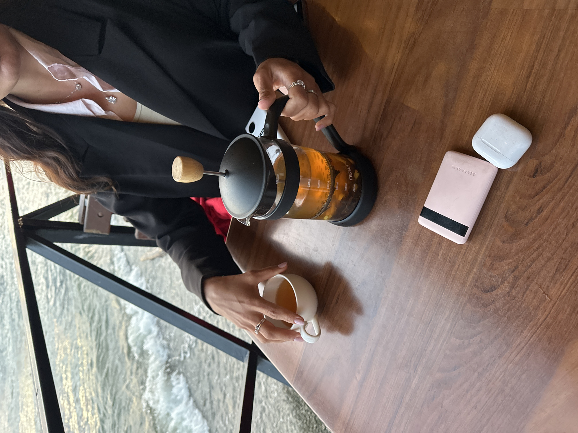 An underexposed, eye-level shot shows a person's hands on a wooden table beside a body of water. The person is wearing a black blazer over a light pink shirt and a white undershirt. They have delicate rings on their fingers and delicate necklaces. Their right hand is on the handle of a black French press containing amber liquid and tea leaves. Their left hand is reaching for a small white teacup filled with amber liquid. On the table, there is a pink portable charger and a white earbud charging case. The background shows a railing and the water. This scene appears to be in Petropavlovsk-Kamchatskiy, Russia, specifically in a cafe with outdoor seating overlooking the Avacha Bay, likely during late afternoon or early evening given the lighting.