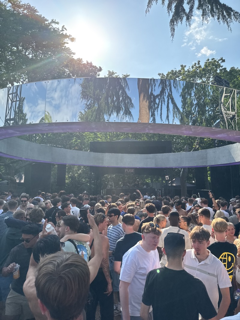 The image shows a large crowd of people gathered at an outdoor event. Above them is a circular structure, partially reflective, with "FUSE" printed on it. The environment appears to be a park or green space, with trees visible in the background and reflected in the structure. The location is likely Barking, United Kingdom, based on the context. The people are interacting, some with arms raised, suggesting they are enjoying a performance or music.