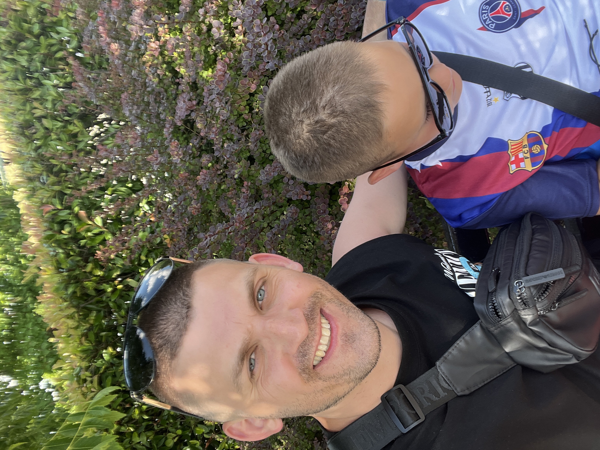 This is a selfie taken outdoors in Ravda, Bulgaria. The image features two individuals, an adult male and a younger male. The adult male is in the foreground, smiling and looking at the camera. He has short hair, blue eyes, and is wearing sunglasses pushed up on his forehead, a black t-shirt, and a black fanny pack. The younger male is to the right of the adult, with his head tilted down and wearing sunglasses. He is wearing a white, blue, and red soccer jersey. They are both seated, with a backdrop of green and red foliage. The lighting suggests it is daytime.