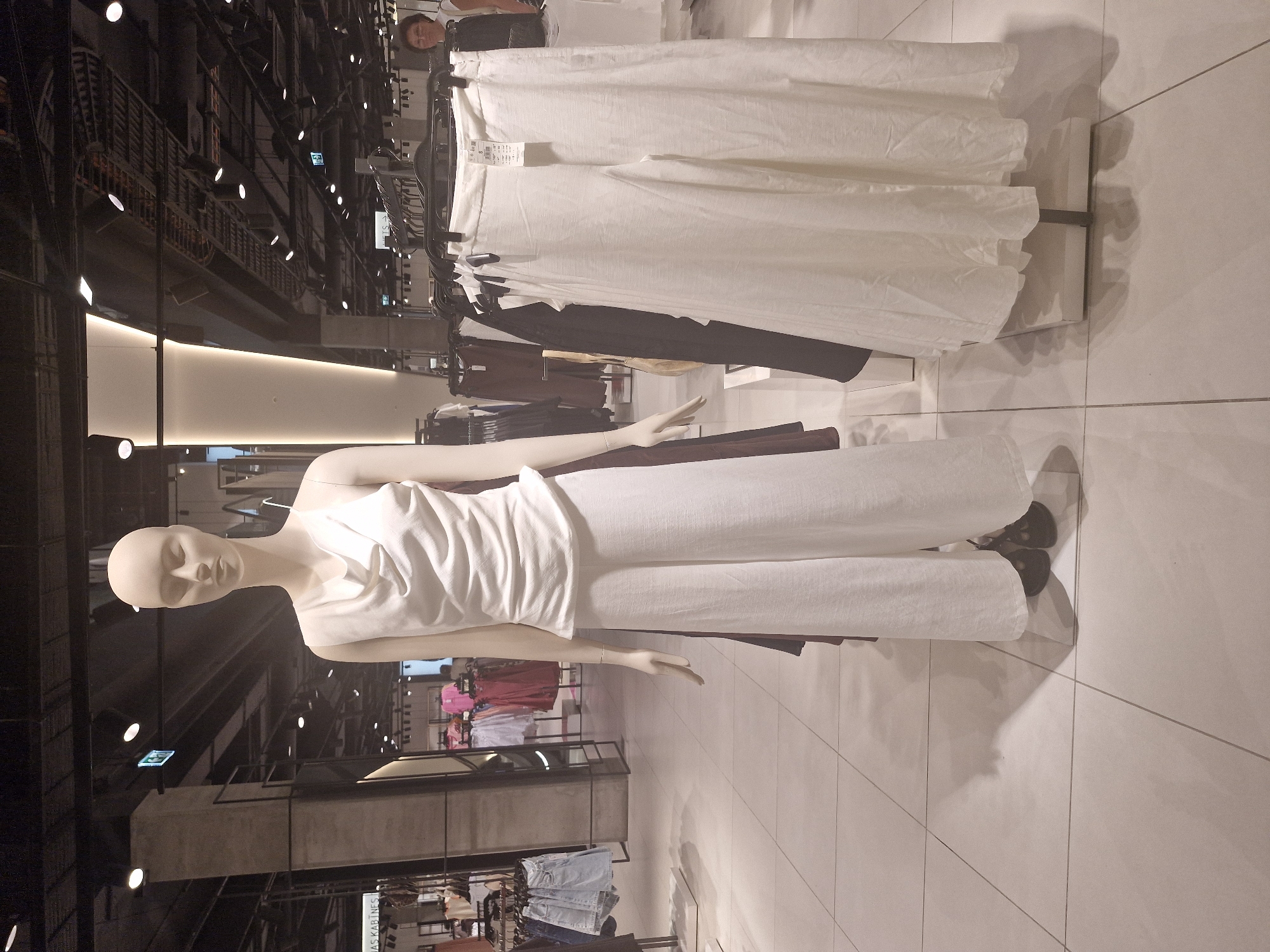 The image displays a clothing store interior, primarily featuring a mannequin modeling an all-white outfit. The mannequin, positioned centrally and oriented vertically (the image is rotated 90 degrees counter-clockwise), has light-toned skin and a smooth, featureless face with closed eyes. It wears a white sleeveless top with a softly draped cowl neck and matching wide-leg trousers, with dark shoes visible beneath the pants. Its arms are held straight down, with hands slightly cupped.
The setting is a modern retail space with a light-colored tiled floor. The ceiling is dark and industrial, with exposed structures, pipes, and numerous bright circular lights casting reflections throughout the store.
In the background, behind the mannequin, several racks of clothing are visible. A prominent rack holds more white garments, likely trousers, similar to those worn by the mannequin. Below these, racks display darker clothing items, including black and brown pants. Further in the background, other clothing racks feature various items, including pink tops and folded stacks of denim jeans.
Notable details include price tags attached to the clothing on the racks. A reflection of a person's head and upper body, appearing to be a woman with dark hair, is visible in the upper right portion of the image, near a clothing rack. An illuminated green exit sign is also reflected in the background. Partial text "AS KABINES" can be seen on a sign in the lower background, likely indicating changing or fitting rooms ("kabines" being a foreign word for cabins/fitting rooms). The overall impression is that of a brightly lit, modern clothing store showcasing a display.