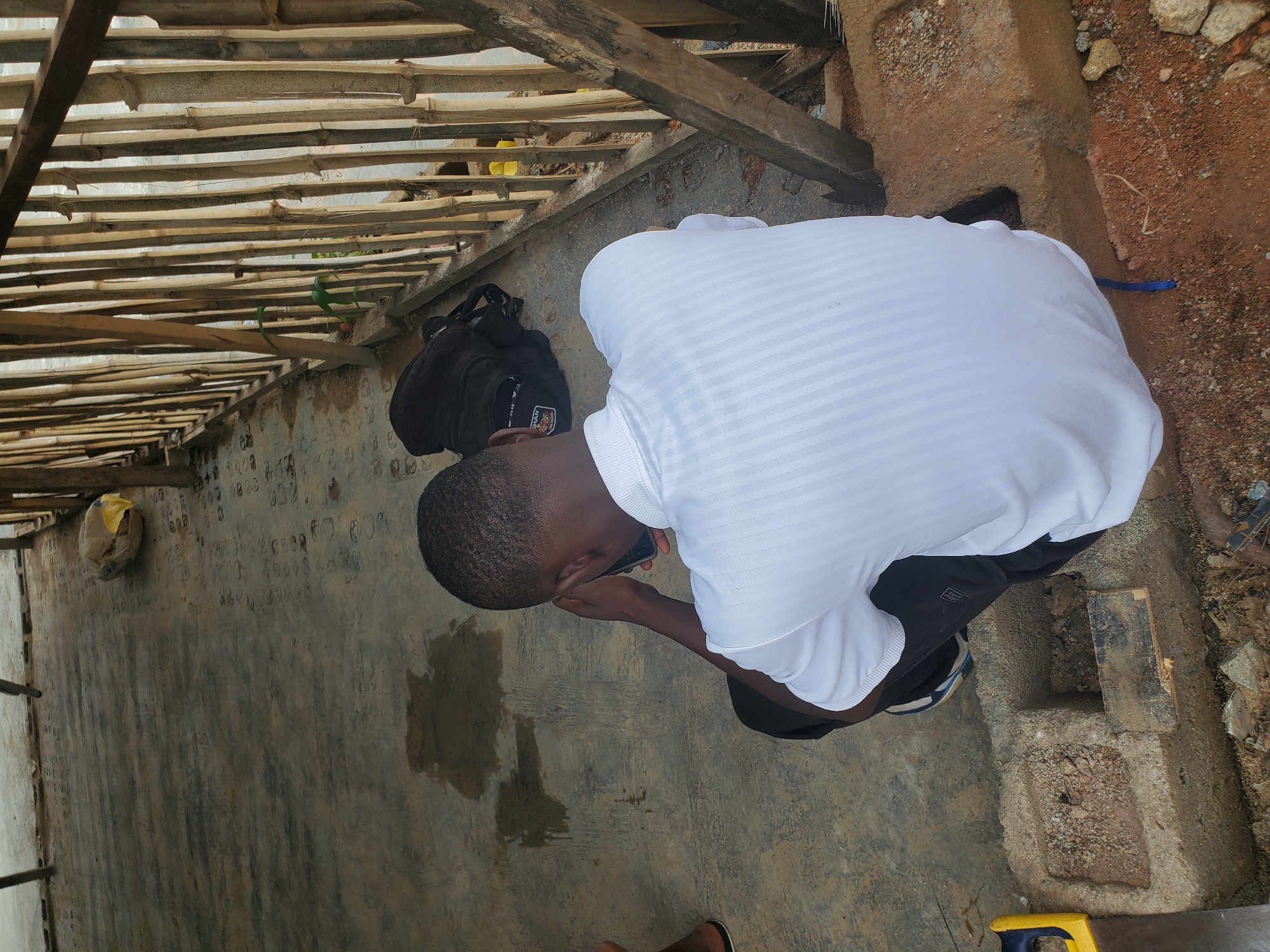 A man is crouched down while using a mobile phone. He is wearing a white shirt and black shorts. A black backpack is near him on the concrete floor of a building under construction, with a bamboo framework visible above. The location appears to be a building site in Jos, Nigeria, as suggested by the environment, building style and the appearance of the individual. Construction materials are present.