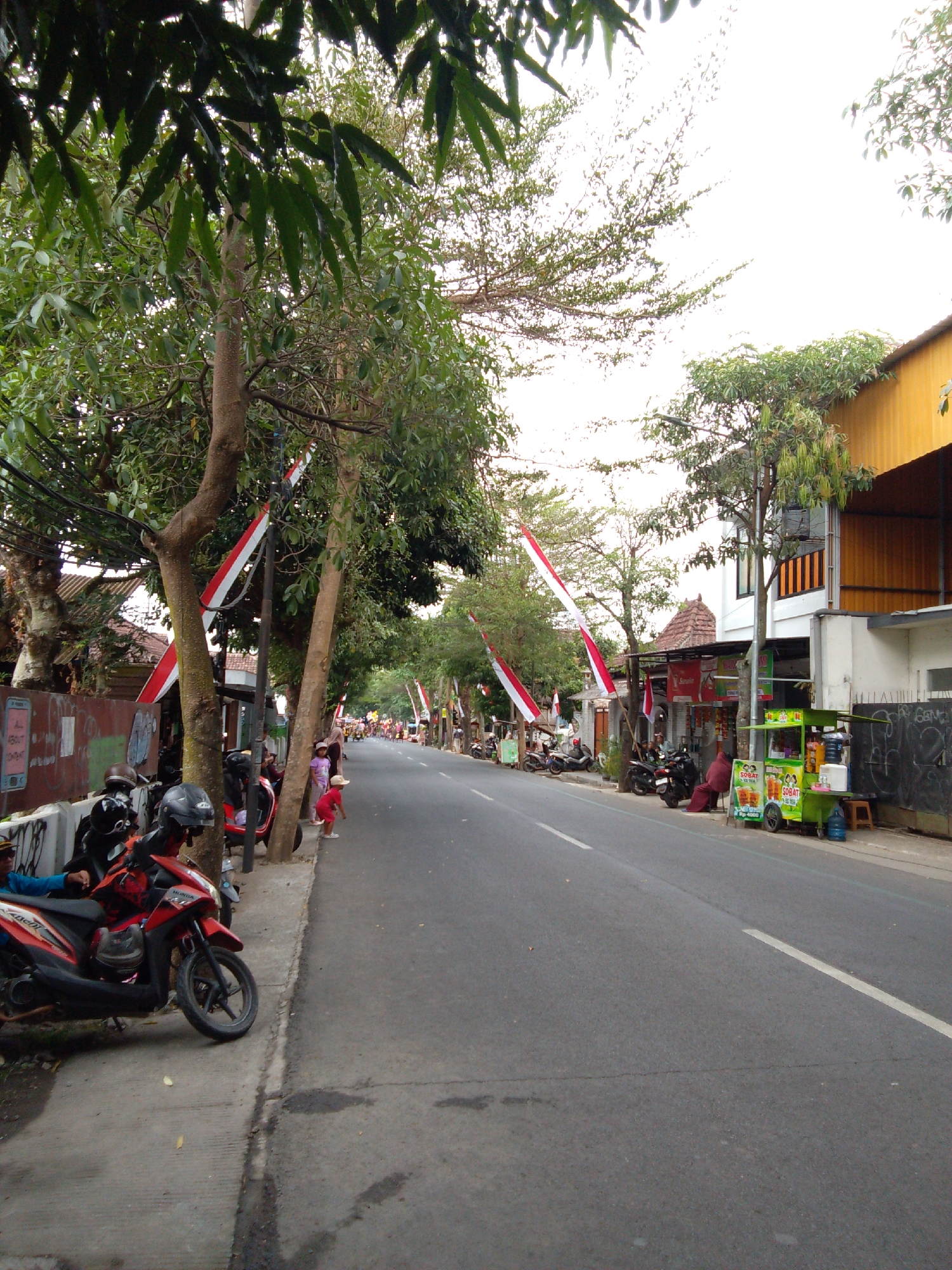 The image depicts a street scene in Trenggalek, Indonesia, likely during a festive occasion given the red and white Indonesian flags decorating the trees and poles lining the road. The street is paved and appears to be a two-lane road with a white line marking the center.
On the left side of the street, a sidewalk is visible. Several motorcycles are parked along this sidewalk, including a red scooter in the foreground. Some people are gathered near the motorcycles, including a person wearing a blue shirt and another wearing a blue jacket. Further down the sidewalk, there are more motorcycles and some individuals standing or walking. The background on the left shows buildings with tiled roofs and some murals on a wall.
The right side of the street features several buildings, including a two-story building with a yellow awning. In front of one of the buildings, there is a green beverage cart selling drinks. A person in a dark red outfit is sitting next to the cart. Further down the street, more flags are visible, suggesting a continuation of the festive decorations. Trees with green foliage are scattered throughout the scene, providing shade. The sky is overcast.
The overall impression is a lively street scene in a residential or commercial area of Trenggalek, adorned for a celebration.