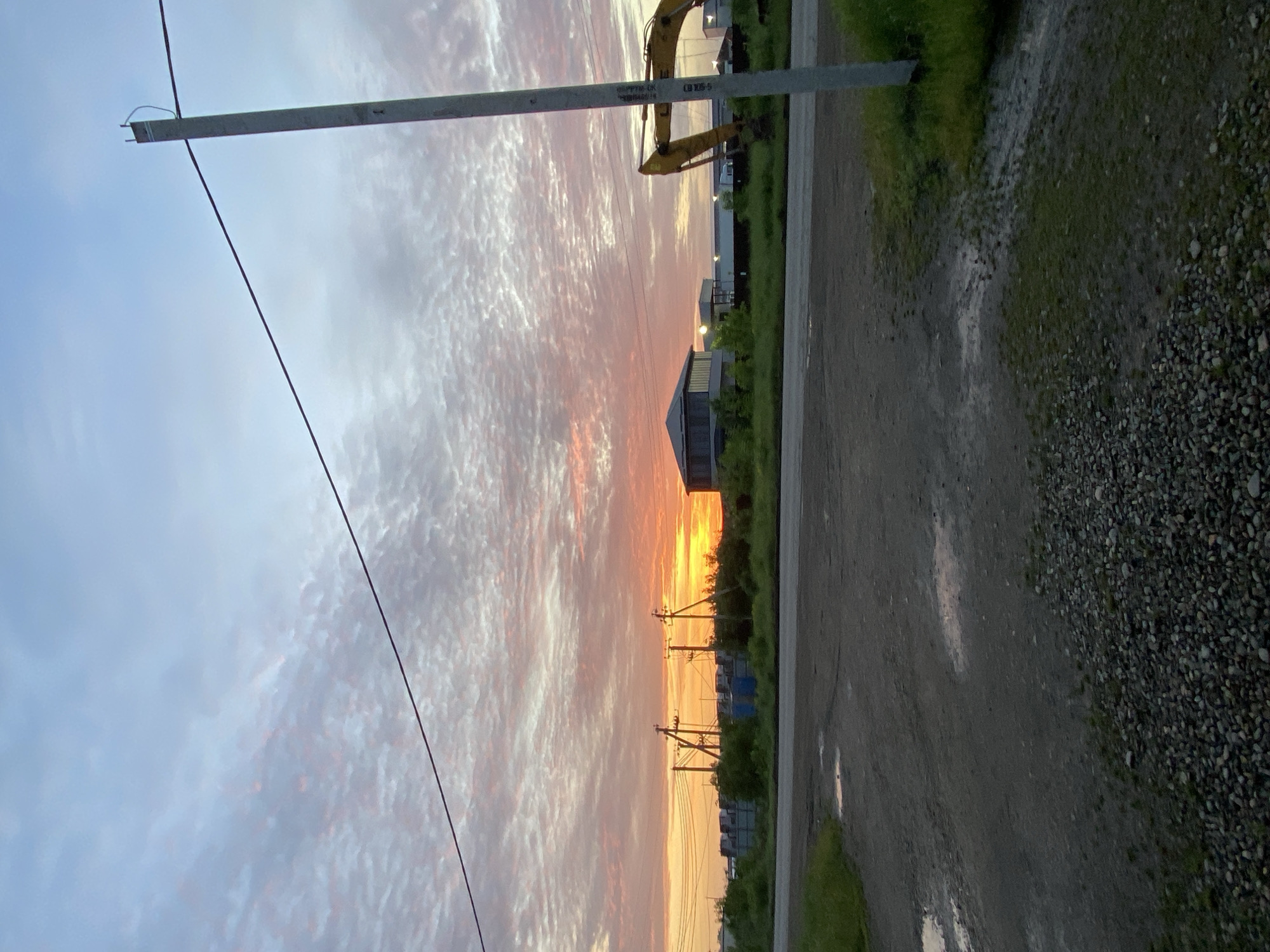 The image depicts a landscape at sunset in what appears to be a rural or semi-rural area in Neftyanik, Russia. The sky is filled with a vibrant mix of colors, ranging from orange and yellow near the horizon to lighter shades of pink and grey higher up. Power lines and utility poles stretch across the sky. On the right side of the image, a road or path is visible, alongside some green vegetation. Several buildings, including what appears to be an excavator, are also present. The overall setting suggests a location on the outskirts of the city.