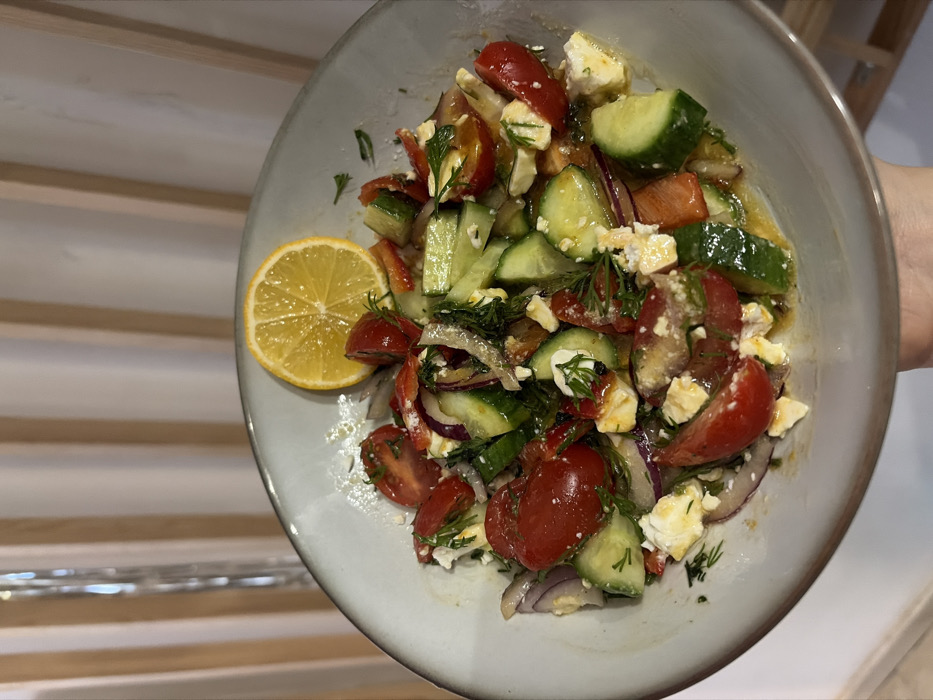 A light gray ceramic bowl containing a fresh salad is held by a person's hand against a background of vertical beige and white stripes. The salad comprises halved red cherry tomatoes, sliced green cucumbers, thinly sliced red onion, crumbled white feta cheese, and chopped green dill, all coated in a visible oily dressing pooling at the bottom of the bowl. A single, thin yellow lemon slice rests on the rim of the bowl, partially submerged in the salad. The background appears to be an interior setting with vertical elements, possibly blinds or wall paneling. The location is Unknown, Unknown.