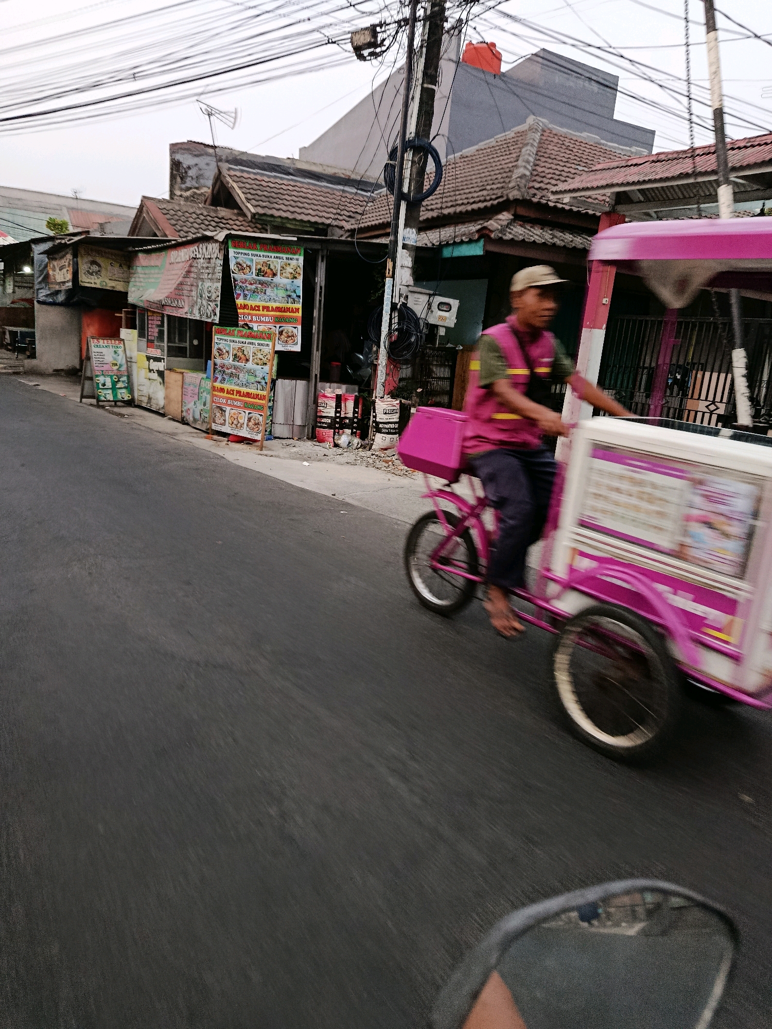 An outdoor, slightly blurred, medium shot captures a man riding a pink ice cream cart down a paved street in Bekasi, Indonesia. The man, wearing a tan cap and a bright pink vest over a green shirt and dark pants, is pedaling the cart, which has a white and pink body with graphics. The cart is blurred due to motion, as are the wheels of the vehicle. The street is asphalt, and on the left side of the image, there are several shops with awnings and posters advertising food and drinks. Overhead, a network of electrical wires crisscrosses the sky, which is overcast. The buildings appear to be single-story structures with tiled roofs. The overall scene depicts a typical street view in a commercial area of the city.