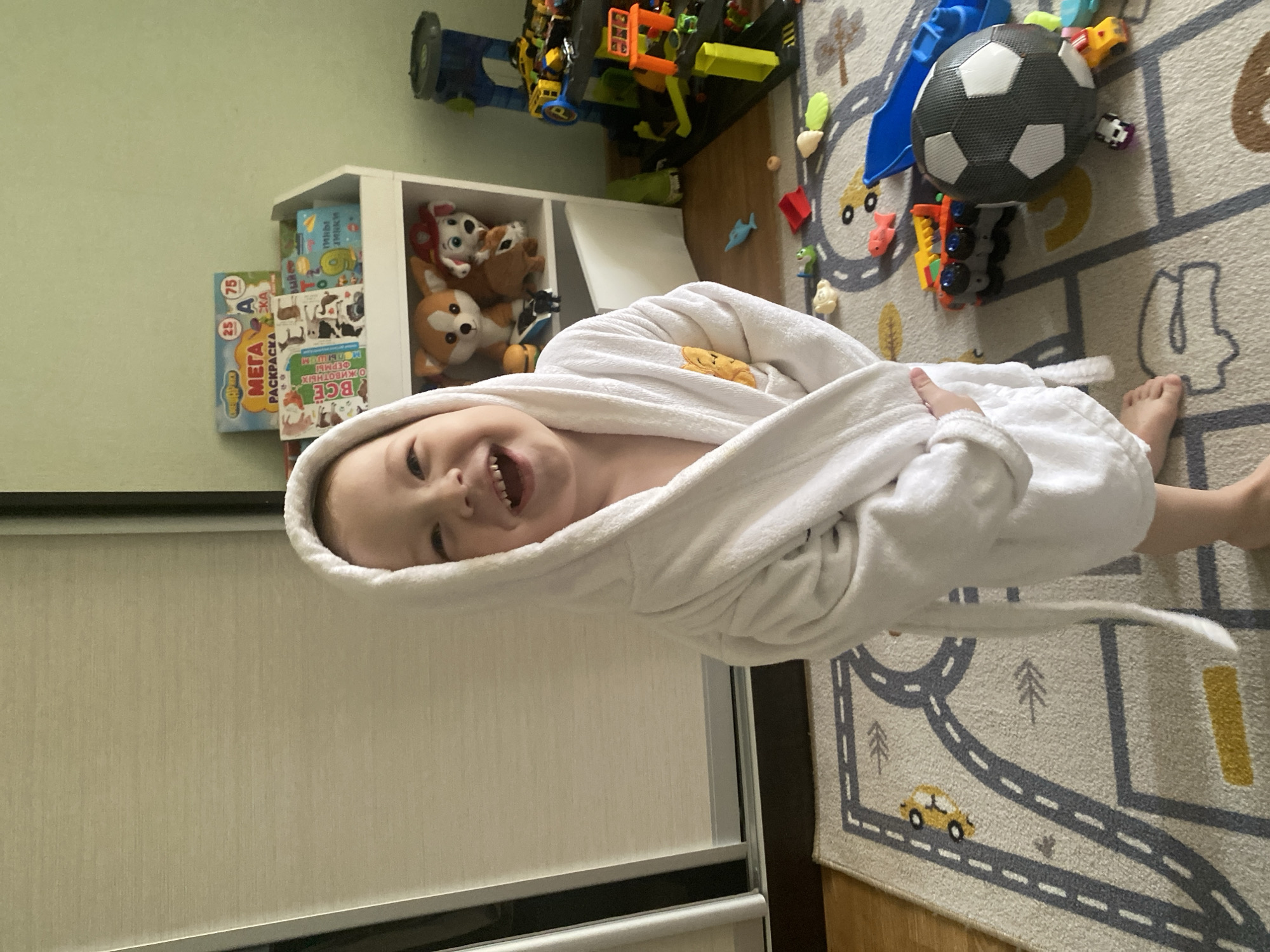 A young child wearing a white hooded bathrobe is smiling and looking towards the camera. The child is standing on a rug that has road patterns and toy cars printed on it. Behind the child is a white bookshelf with toys and books. Above the bookshelf are more toys. A soccer ball is on the rug. The location appears to be indoors, likely a child's playroom. There is no indication of this being Sochi, Russia.