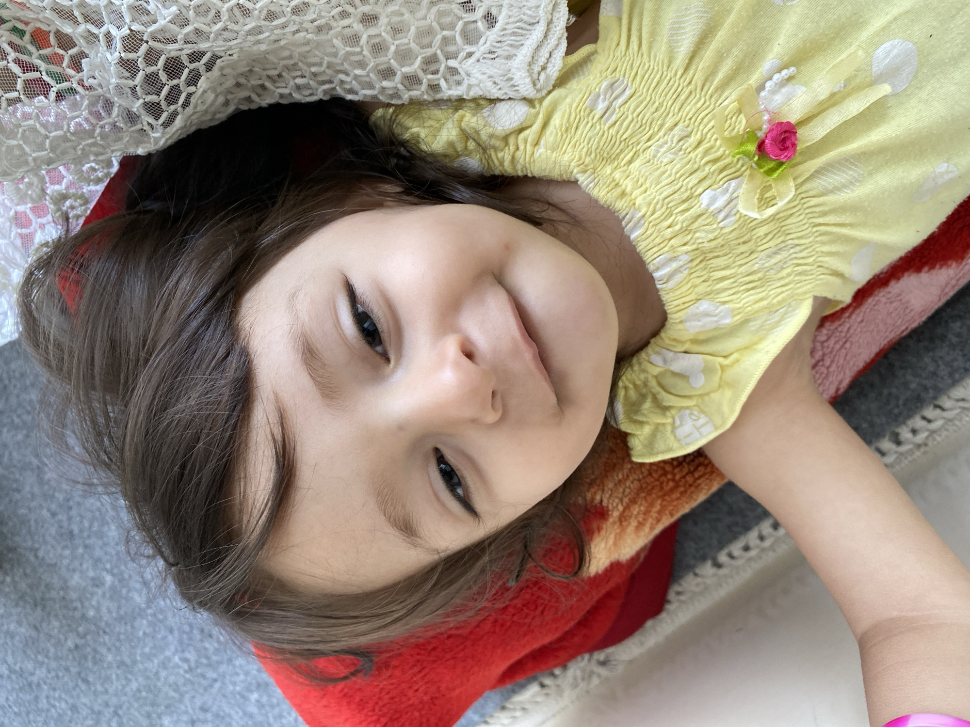 This close-up image features a young child, likely a girl, with dark brown hair and dark eyes, lying down and looking directly at the camera. Her head is tilted slightly, and she has a gentle, playful smile with her mouth slightly open.
The child is wearing a light yellow top or dress adorned with white polka dots. The upper part of the garment features a ruched or smocked texture. A decorative detail, including a small pink fabric rose, a green leaf, and a short string of tiny white beads, is attached to the shoulder area of her top.
A piece of white, intricately crocheted or lacy fabric partially covers the upper left portion of her head and extends above her, suggesting she might be under a decorative net or canopy. Her head rests on a soft, red blanket or cushion, which is also visible beneath her body. Hints of other warm colors, possibly orange or brown, are visible at the edges of this bedding. To the left of the red blanket, a gray fabric or carpet is visible, indicating an indoor setting.
The lighting is soft and even, characteristic of an indoor daytime scene. The child's expression conveys innocence and a friendly, engaging demeanor. There is no visible text within the image, and no specific location cues for Kandahar, Afghanistan, are present in the visible environment.