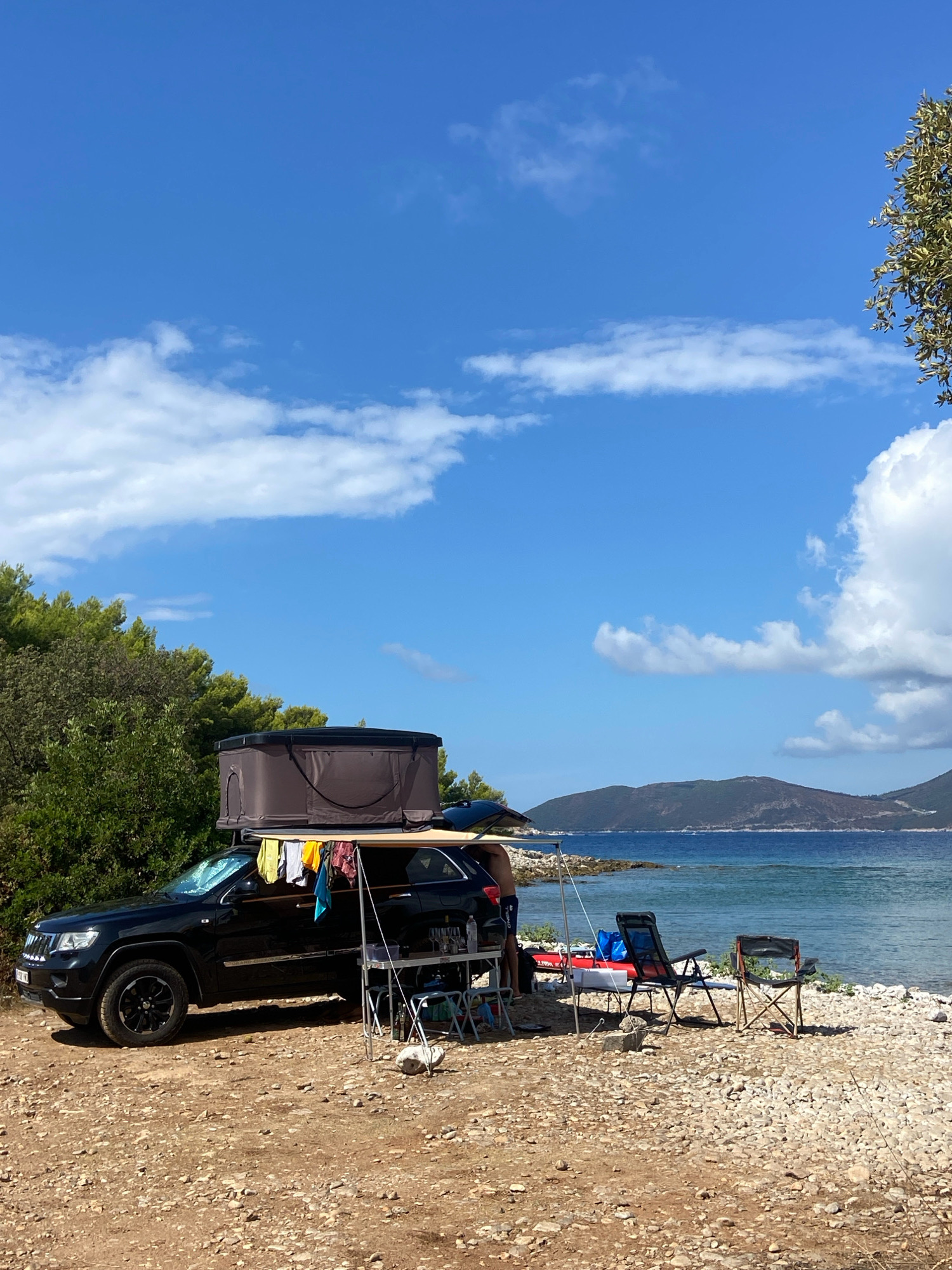 A black Jeep Grand Cherokee with a rooftop tent is parked on a rocky beach next to the Adriatic Sea in Luštica, Montenegro. The vehicle is accessorized for camping, with clothes hanging to dry from a rack attached to the side and a portable table set up with chairs. The water is a clear blue, and the sky is bright with scattered white clouds. In the background, lush green hills rise from the sea. The scene depicts a serene outdoor recreational setting.