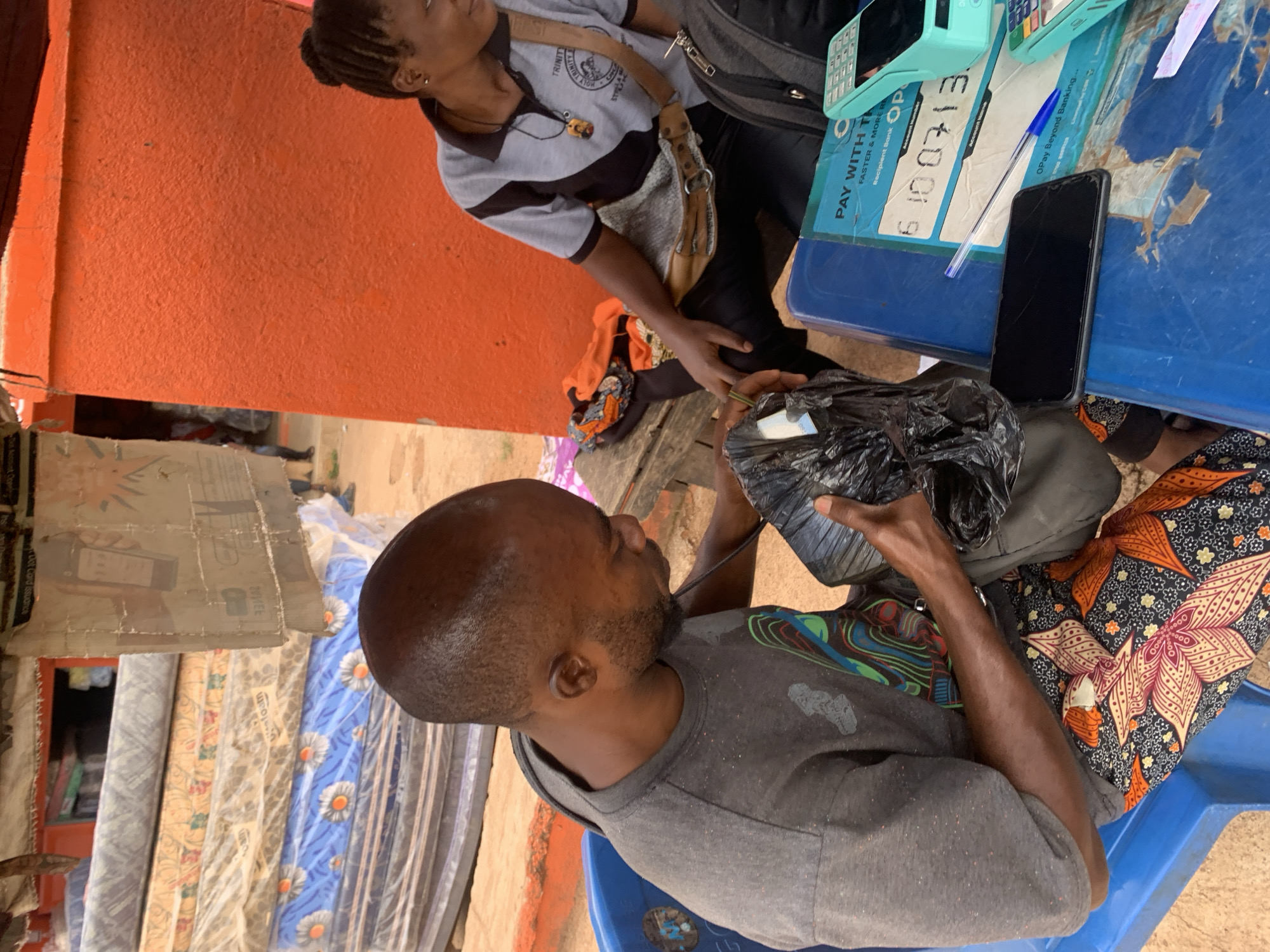 This image captures a daytime scene in what appears to be an outdoor or semi-outdoor commercial setting, possibly a market stall or small shop in Jos, Nigeria. Two individuals are engaged in a transaction.
In the foreground, a man with a beard and short hair is seated, wearing a grey t-shirt with a colorful design. He is looking down and holding a black plastic bag. Partially visible in the background, a woman, likely a vendor, is also seated. She is wearing a grey and black polo shirt with "TRINITY" visible on it, and a brown bag is strapped across her body.
On a blue plastic table between them, several items related to a financial transaction are present. These include two light-blue payment (POS) terminals, a black smartphone with a cracked screen, and a blue pen. A blue document, associated with "OPay Beyond Banking", is also on the table, displaying text like "PAY WITH T", "Recipient Bank OPay", and an account number "6100713". This setup strongly suggests a mobile money payment or withdrawal.
The background features an orange-red wall behind the woman. To the man's left, several rolled-up and stacked mattresses, some with patterned covers, are visible, indicating that the stall might sell bedding. Text like "じゃっfoam" (possibly "Vitafoam") is discernable on one of the mattress covers. The overall scene reflects a common commercial interaction in a local marketplace environment.