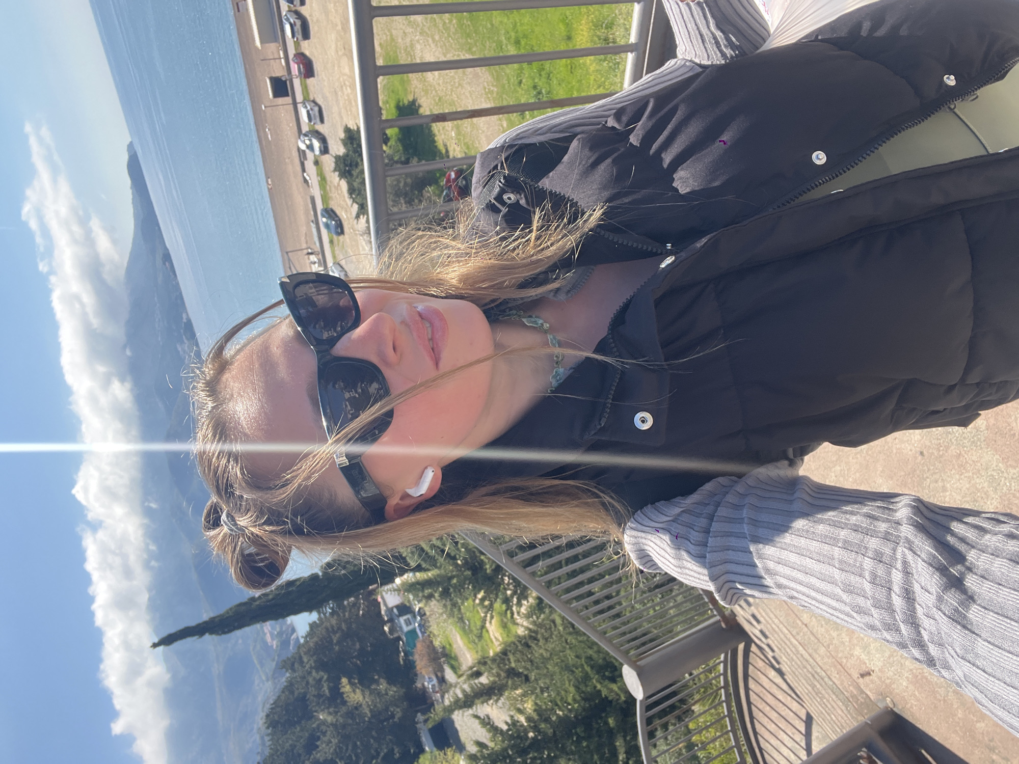 A selfie was taken on an elevated outdoor platform, possibly a balcony or scenic overlook. The subject is a young fair-skinned woman with light brown hair worn in a double bun. She is wearing sunglasses, a black puffer jacket, a grey ribbed long-sleeved shirt, and what appears to be a light green top underneath. She has an earbud in her left ear and is wearing a beaded necklace. The background features a view that includes the Adriatic Sea, a beach with parked cars, and a mountain range. Green vegetation and a slender tree are visible in the foreground. The location is likely the city of Boreti, Montenegro, given the landscape features.