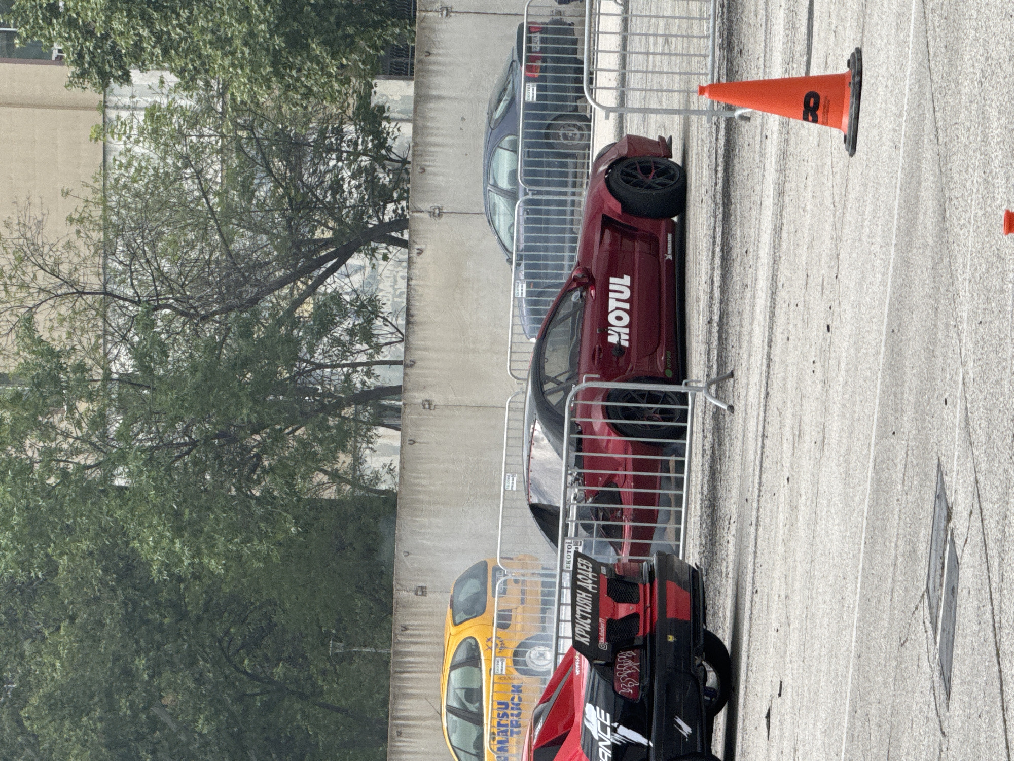 The image shows a section of a parking lot in Plovdiv, Bulgaria, adjacent to a road. Visible are four vehicles parked alongside a metal barrier. From left to right, there is a black and red car with “MATSU TRUCK” written on the side, followed by a yellow car of the same brand. Next is a red car with a white “MOTUL” logo, and then a dark blue or black sedan. An orange traffic cone with the number "8" is visible on the road surface. A tree and the side of a building are on the left. The parking area appears to be marked with painted lines on the asphalt surface.