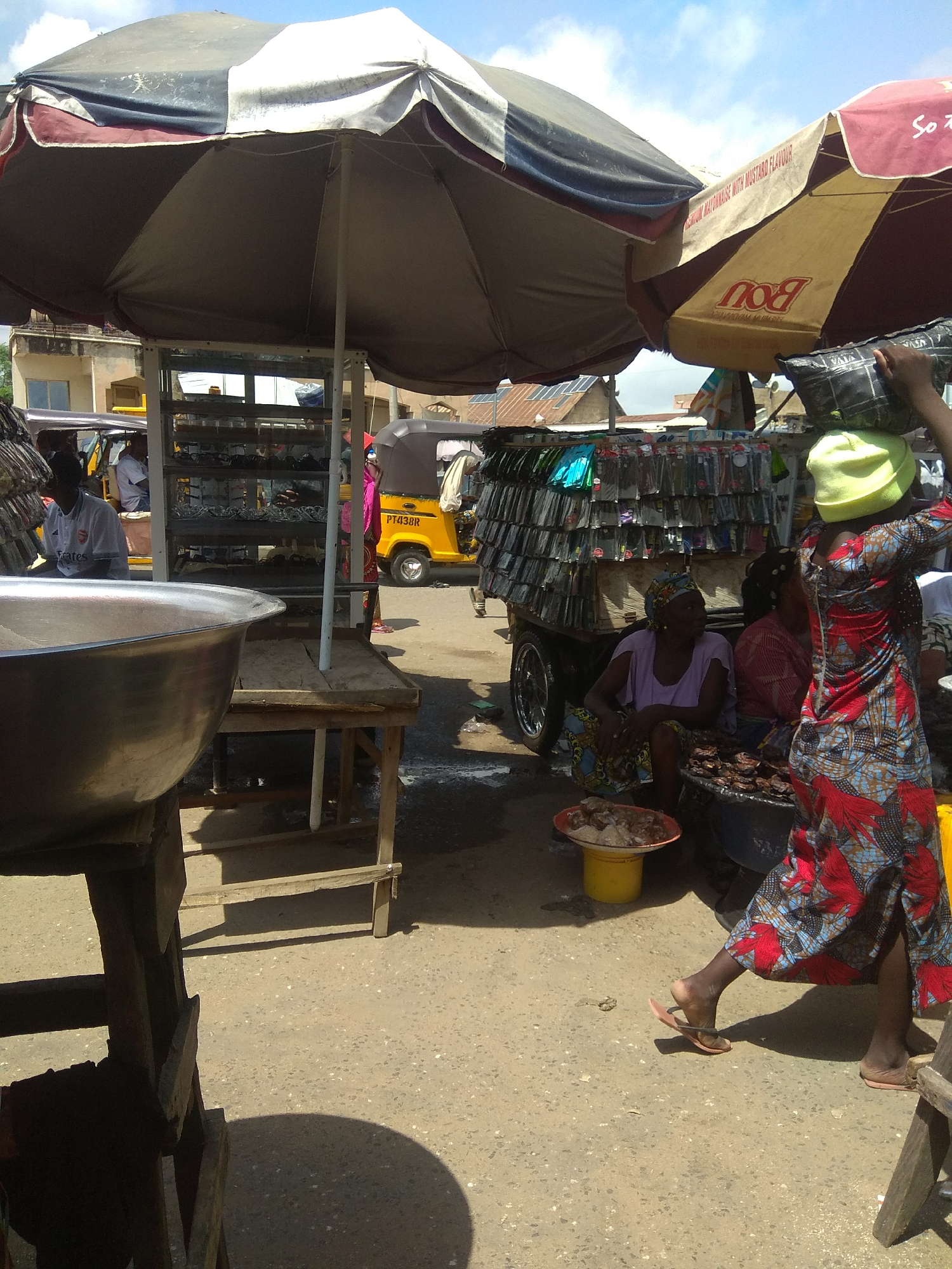 This media file depicts a bustling outdoor market scene in Jalingo, Nigeria. The primary subjects are several people, likely vendors and shoppers, engaged in their daily activities under the shade of large umbrellas. In the foreground, a woman in a vibrantly patterned red and blue dress carries a load on her head, wearing a yellow head covering and simple flip-flops. Beside her, another woman in a light purple top and a patterned headscarf is seated, possibly attending to a stall. A large metal bowl is prominently displayed on a wooden stand to the left.
Behind them, a mobile stall is laden with numerous items, possibly phone accessories, displayed in clear packaging. A yellow auto-rickshaw, a common mode of transport in the region, is visible in the background, with a person seated inside. Other market stalls and structures can be seen, suggesting a lively commercial environment. The ground is a dusty, light-colored surface, characteristic of outdoor markets.
The lighting suggests it is daytime, with strong shadows indicating a sunny but not necessarily midday sun. The presence of umbrellas signifies an effort to provide shade from the sun. The overall atmosphere appears to be one of commerce and daily life. No discernible emotions are overtly expressed by the individuals, but the setting suggests activity and potentially a moderate level of heat.
There is some visible text on the umbrellas and the auto-rickshaw. One umbrella has "Bou" in large letters, and another has text that includes "PREMIUM MAYONNAISE WITH MUSTARD FLAVOUR". The auto-rickshaw has a license plate that reads "PT438R".